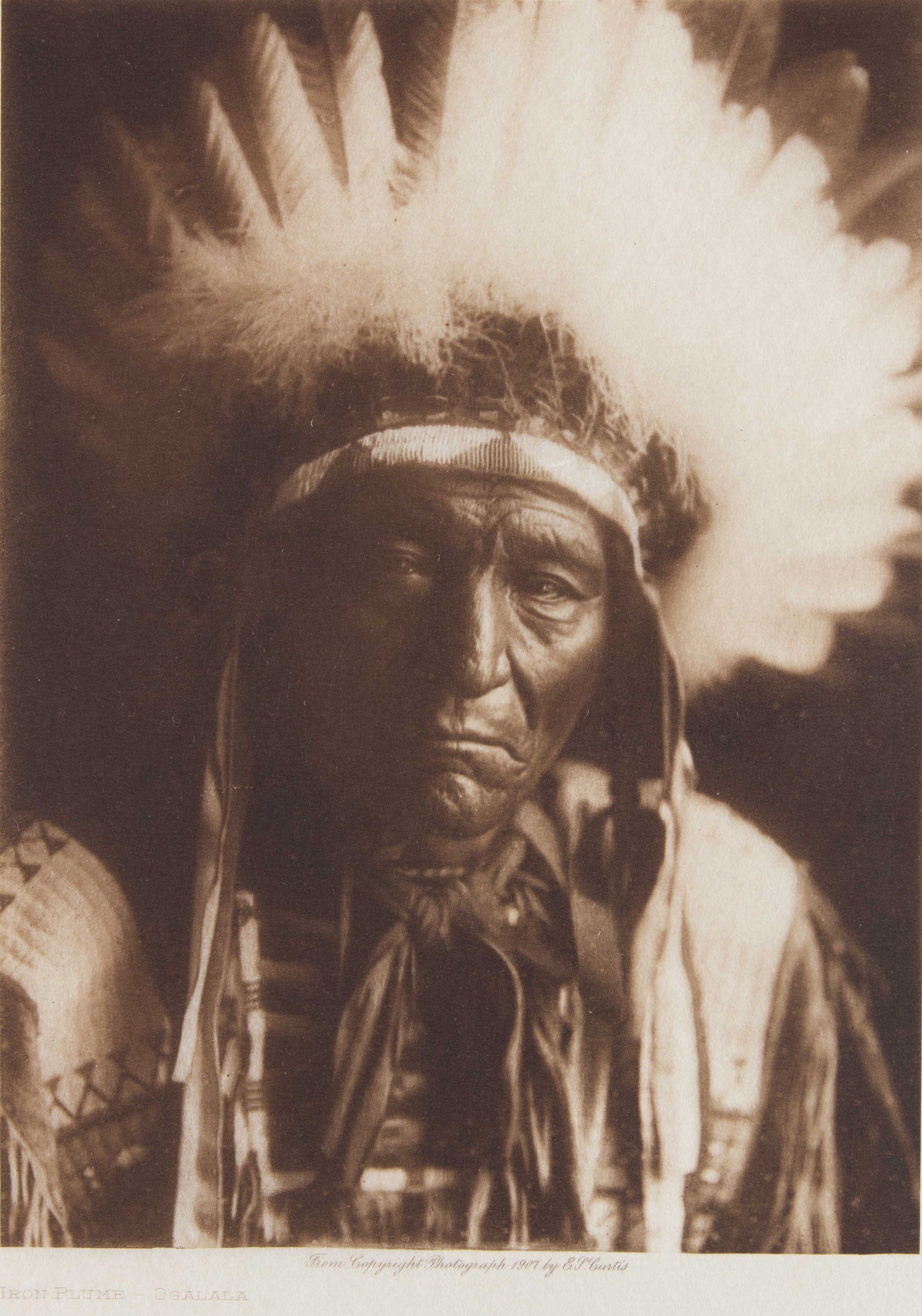 Studio facial portrait is of an older Ogalala man, shown from the chest up, facing forward. The man wears a hide shirt decorated with beadwork panels of geometric designs including triangular motifs on a white background (over figure's proper right shoulder). He also wears a full eagle feather headdress with a beadwork rim/edging (inverting white and colored triangle design).