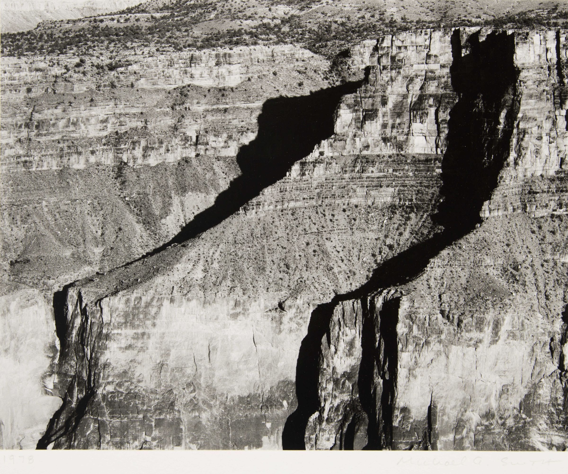 Black and white still photograph of Toroweap, Arizona. Landscape is of an open, aerial side view of a massive rock canyon wall. The wall has three flat facing sides with two jagged, receding formations/crevices (over from the right side of the viewing plane). Narrow straight horizontal rock layers are evident across top of canyon rock wall.
