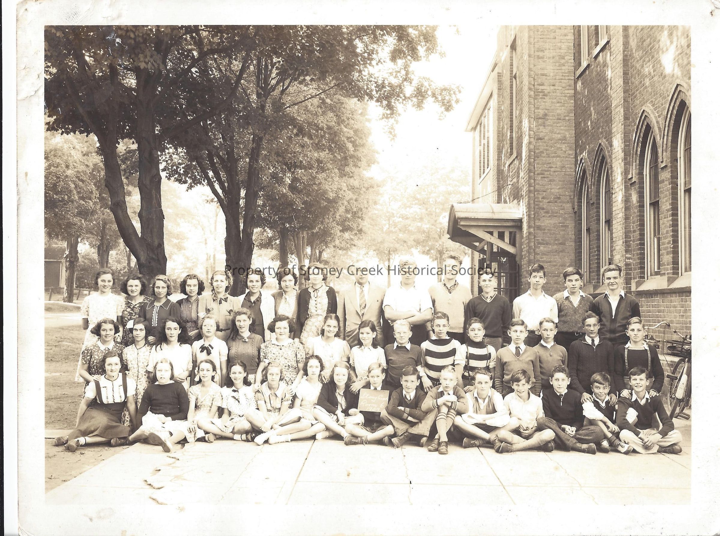 The image portrays a group of approximately 45 people, likely a school class, posing together for a photo outdoors in front of a large building with prominent brickwork and arched windows. The group, comprising of various ages and arranged in four rows, includes mostly younger individuals and a few adults, possibly teachers. Most are dressed in typical mid-20th-century attire, making it likely that this photo was taken in that era. The setting is serene, framed by lush trees and a clear pathway, suggesting a close-knit academic or community event captured on a sunny day.