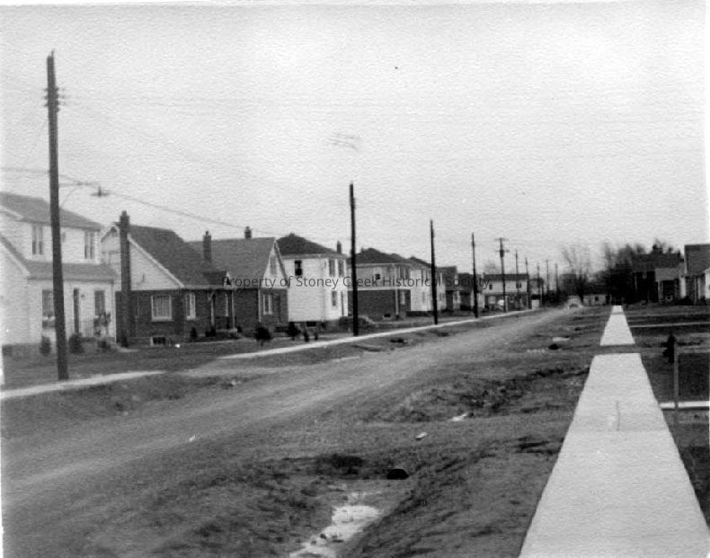 The image shows a suburban street, depicting Randall Ave in Stoney Creek from 1950. It features a row of modest, detached houses with varied facades, neatly spaced along a straight, unpaved road. Sidewalks are visible, but the road surface appears rough and unfinished, with some puddles. Utilities are evident with several poles lining both sides of the street, stretching into the distance under an overcast sky. The neighborhood conveys a quiet, mid-century suburban environment, typical of post-war residential development.