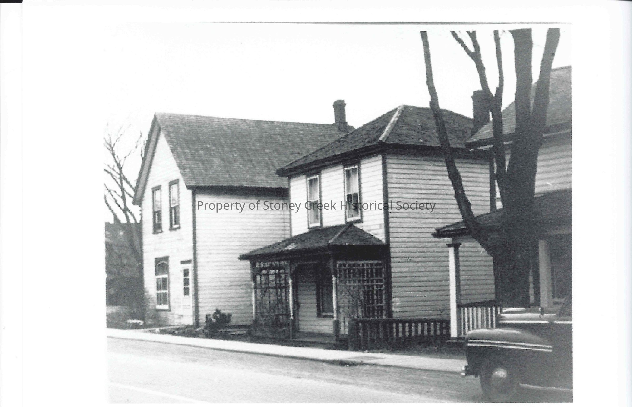 The image captures a historical street scene from downtown Stoney Creek in the 1940s, showing MacDougall's original shop located on the far left. This black-and-white photograph features two prominent buildings with wooden siding and a classic car parked on the street. The architecture suggests a quaint, mid-20th-century small-town vibe, with details like the small porch and awning, characteristic of the period's store fronts.