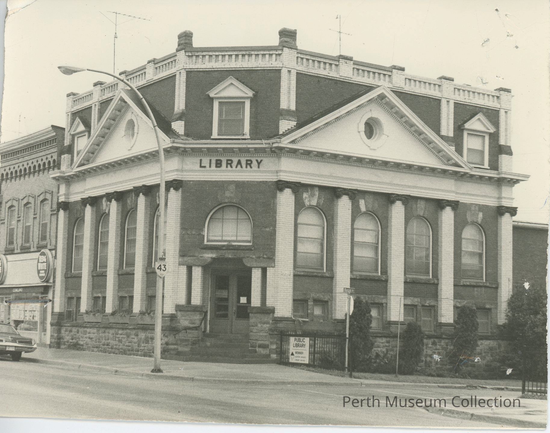 "Perth Library - Carnegie Building": The image depicts an old library building with classic architectural features, such as large arched windows and decorative columns, which dominate the façade. The structure, constructed of brick and stone, showcases an elegant, historical design with intricate detailing around its roof and eaves. A sidewalk runs along the building's perimeter, and a road sign labeled "43" is visible near the entrance, indicating a busy street intersection. Trees are planted nearby, adding a touch of greenery. A visible sign reads "PUBLIC LIBRARY".