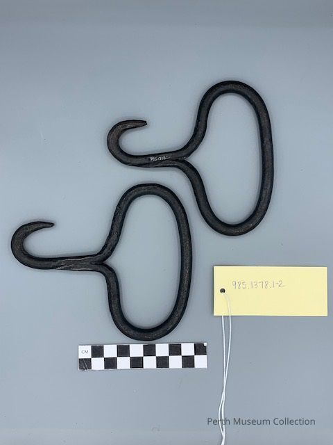 Forged Iron Hooks: The image displays two black, metal devices resembling old-fashioned hooks with looped handles, arranged side by side on a plain gray background. Each hook has a curved, pointed end and a distinctive shape, suggesting a utilitarian purpose. The hooks appear aged, indicating possible historical or archaeological significance, and their placement