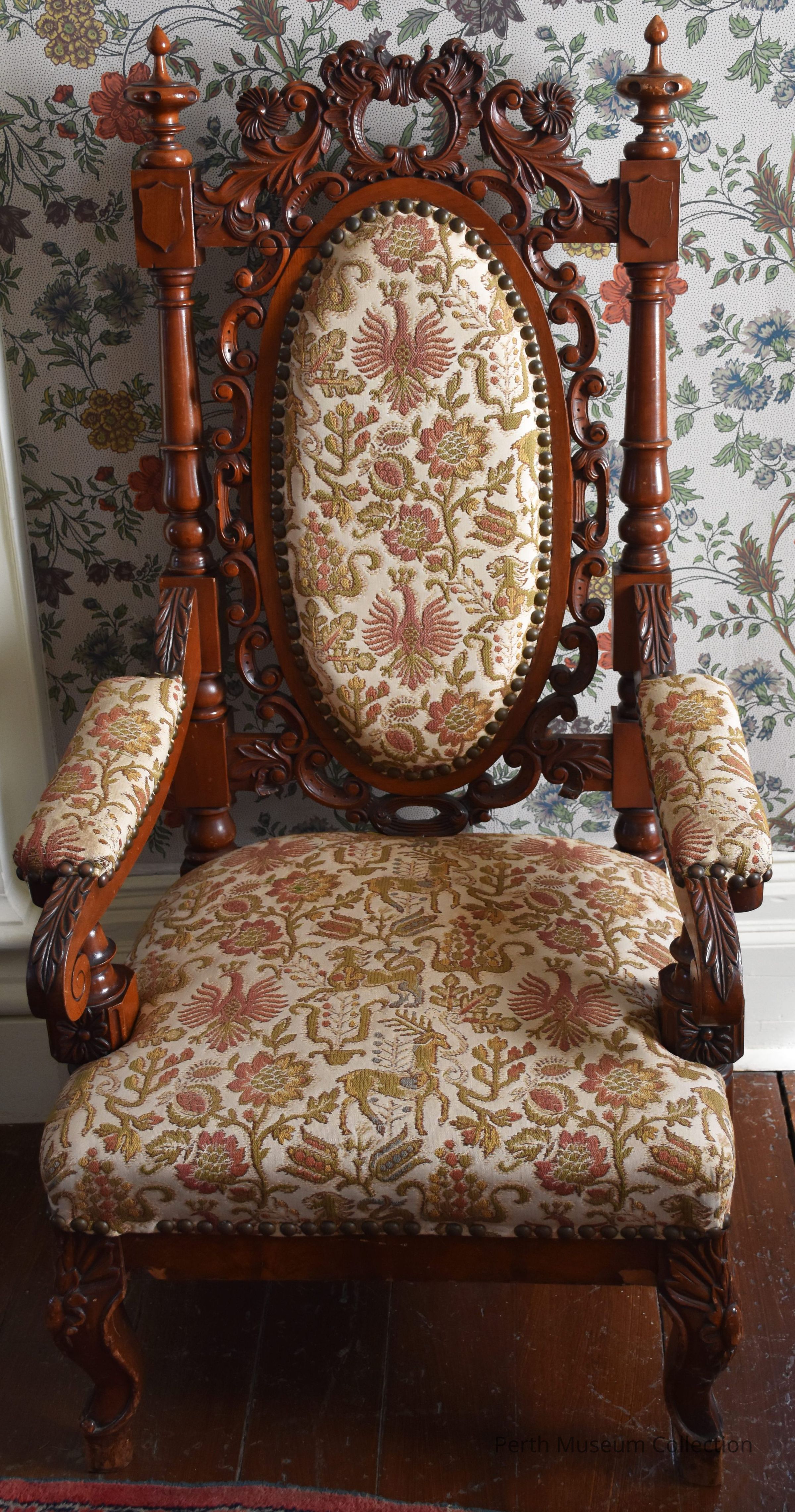 Chair: The image showcases an ornate wooden chair with intricate carvings on its frame, suggesting a classical design. Upholstered with a richly patterned fabric featuring floral and animal motifs in muted tones of red, gold, and green, the chair exudes a historic and luxurious ambiance. It stands on a polished wooden floor, accompanied by wallpaper decorated with floral patterns that complement the fabric. The high-backed chair is flanked by carved wooden armrests, and each detail emphasizes craftsmanship, creating a harmonious blend of elegance and tradition.