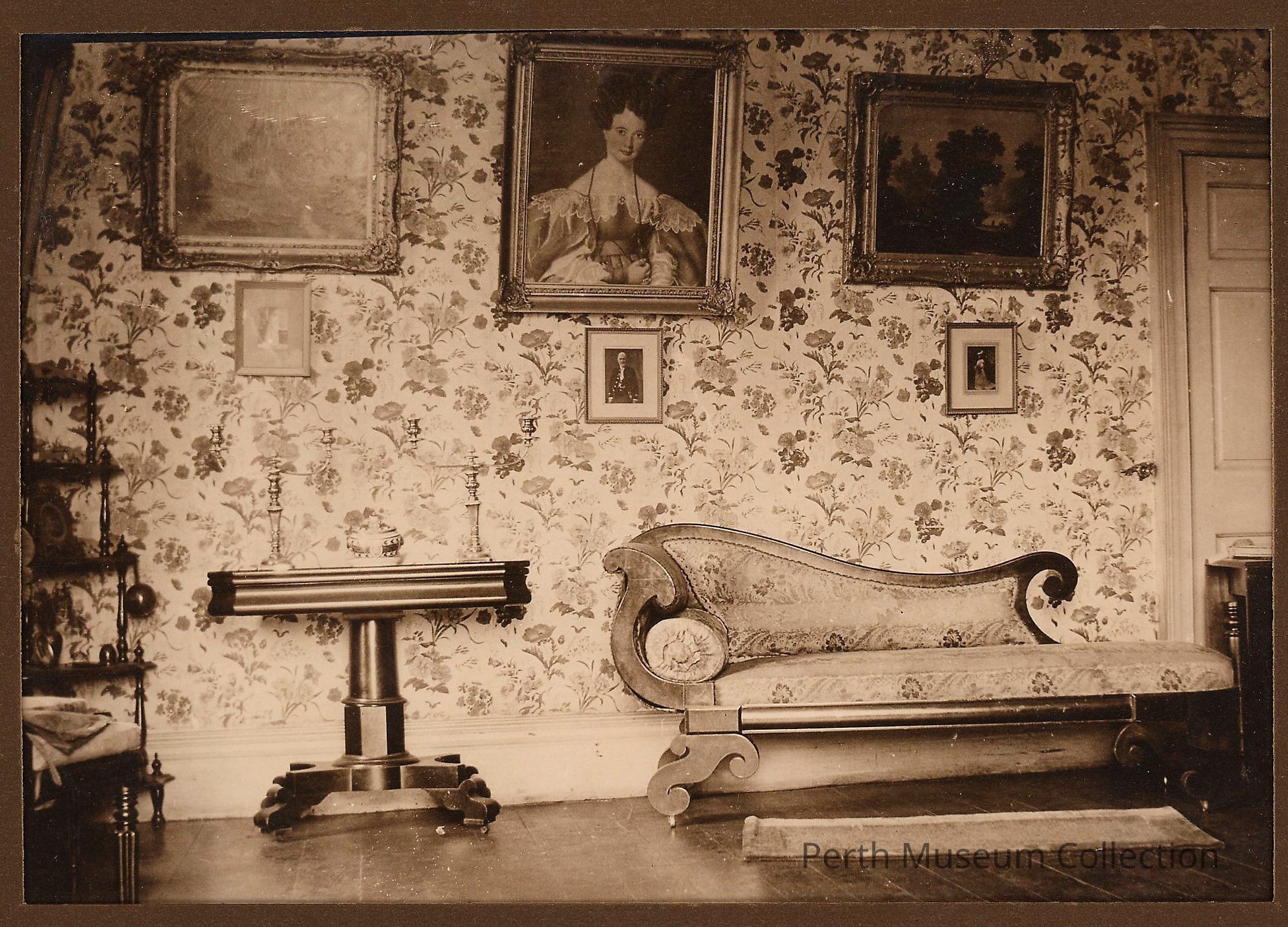 Drawing Room: The image depicts a vintage room with floral wallpaper, adorned with framed artwork, including a large portrait and landscape paintings. A chaise longue with ornate wooden details and a patterned fabric aligns with the right wall, while a polished wooden console table with decorative candlesticks stands to its left. To the side, a corner shelf holds various objects, adding to the room's antique ambiance. The hardwood floor shines beneath a small rug, and a partially visible doorway suggests the room's historical charm.