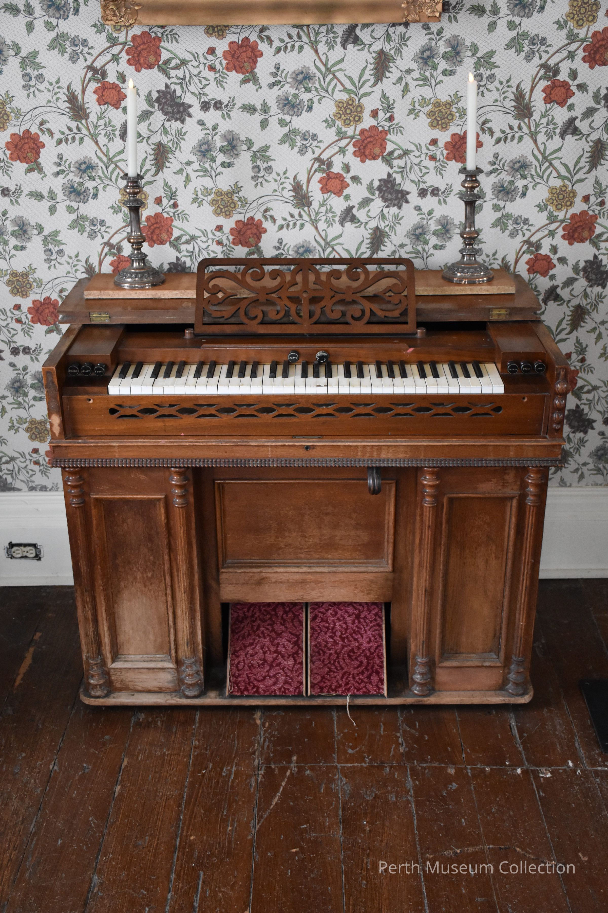 Pump Organ: The image depicts a vintage harmonium with ornate wooden craftsmanship typical of classic furniture design, featuring intricate carvings on the music stand and sides. It rests against a quaint floral wallpaper, providing a charming and nostalgic ambiance. On top of the harmonium sit two silver candlesticks with white candles, alongside a few books, adding to the old-world aesthetic. The overall setting suggests an elegant, historical space where music and classic decor blend harmoniously, likely evoking a sense of serene and antique beauty.