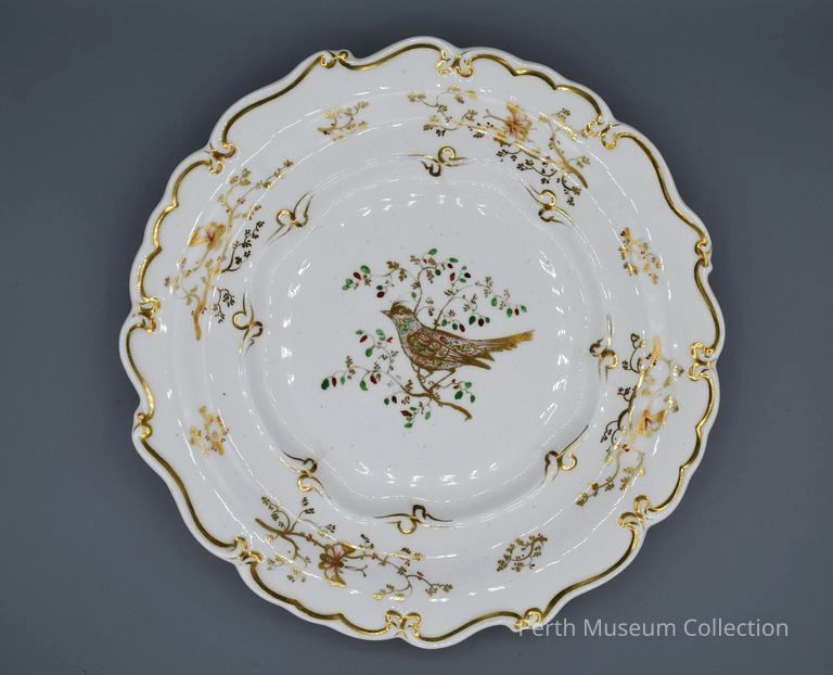 Plate: The image showcases an ornate, round porcelain plate with a beautifully scalloped edge, adorned with a delicate gold trim. At the center of the plate is an intricate design of a bird perched on a branch, with green and gold accents depicting the leaves and flowers. This central motif is surrounded by elegant floral patterns that echo the natural theme and are subtly highlighted with gold detailing. The overall aesthetic of the plate is one of refined elegance, reflecting a harmonious blend of artistic craftsmanship and decorative sophistication.