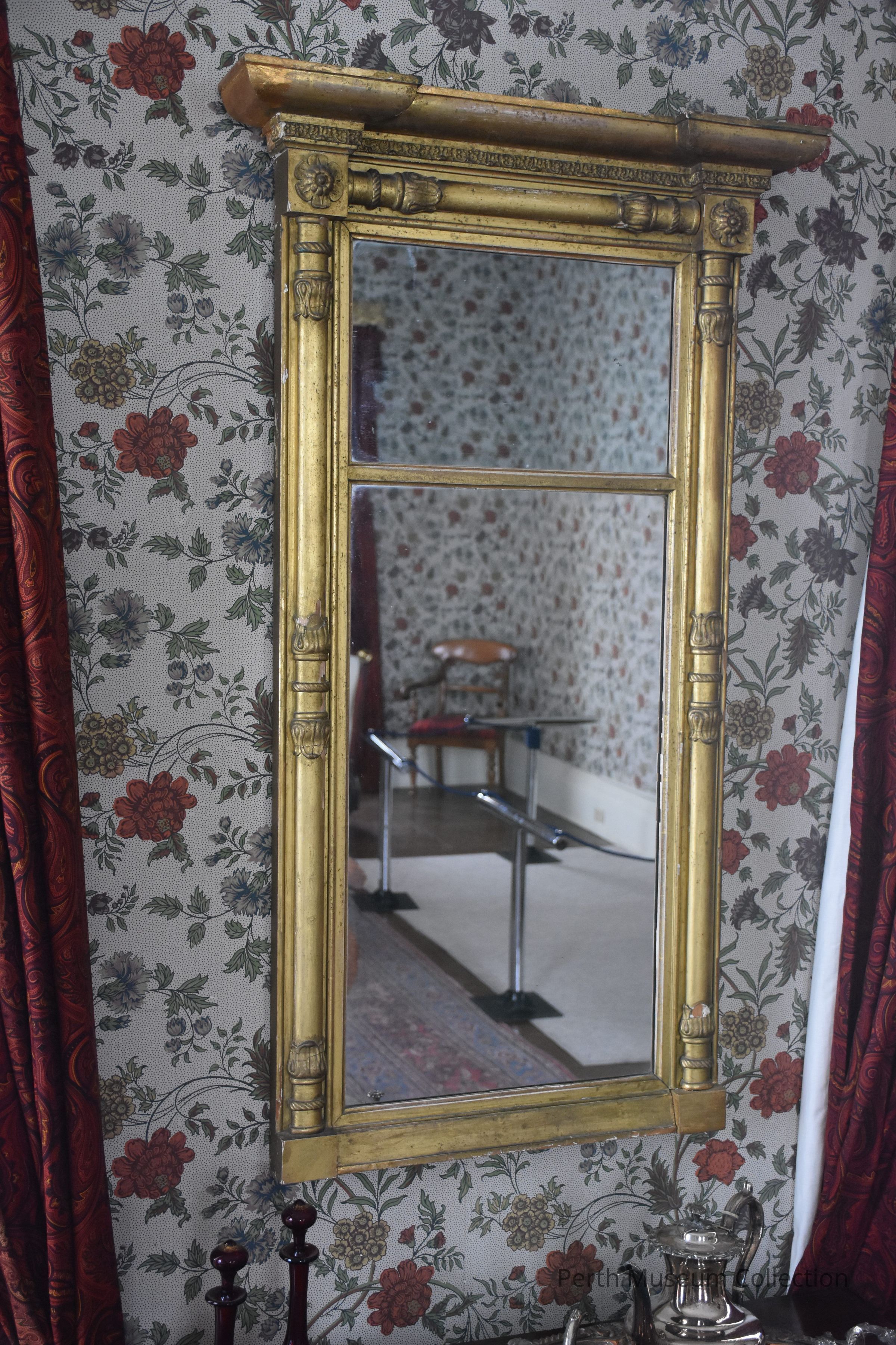 Mirror: A rectangular mirror with an ornate, golden frame hangs on a wall adorned with floral wallpaper featuring red and blue flowers and green leaves. In the reflection, a room with similar wallpaper and a wooden chair can be seen, along with a section of patterned carpet. The rich, red curtains framing part of the mirror add a touch of elegance to the scene. The intricate detailing on the mirror's frame includes small floral and column-like embellishments, enhancing its vintage aesthetic, and the overall setting exudes a classic feel.
