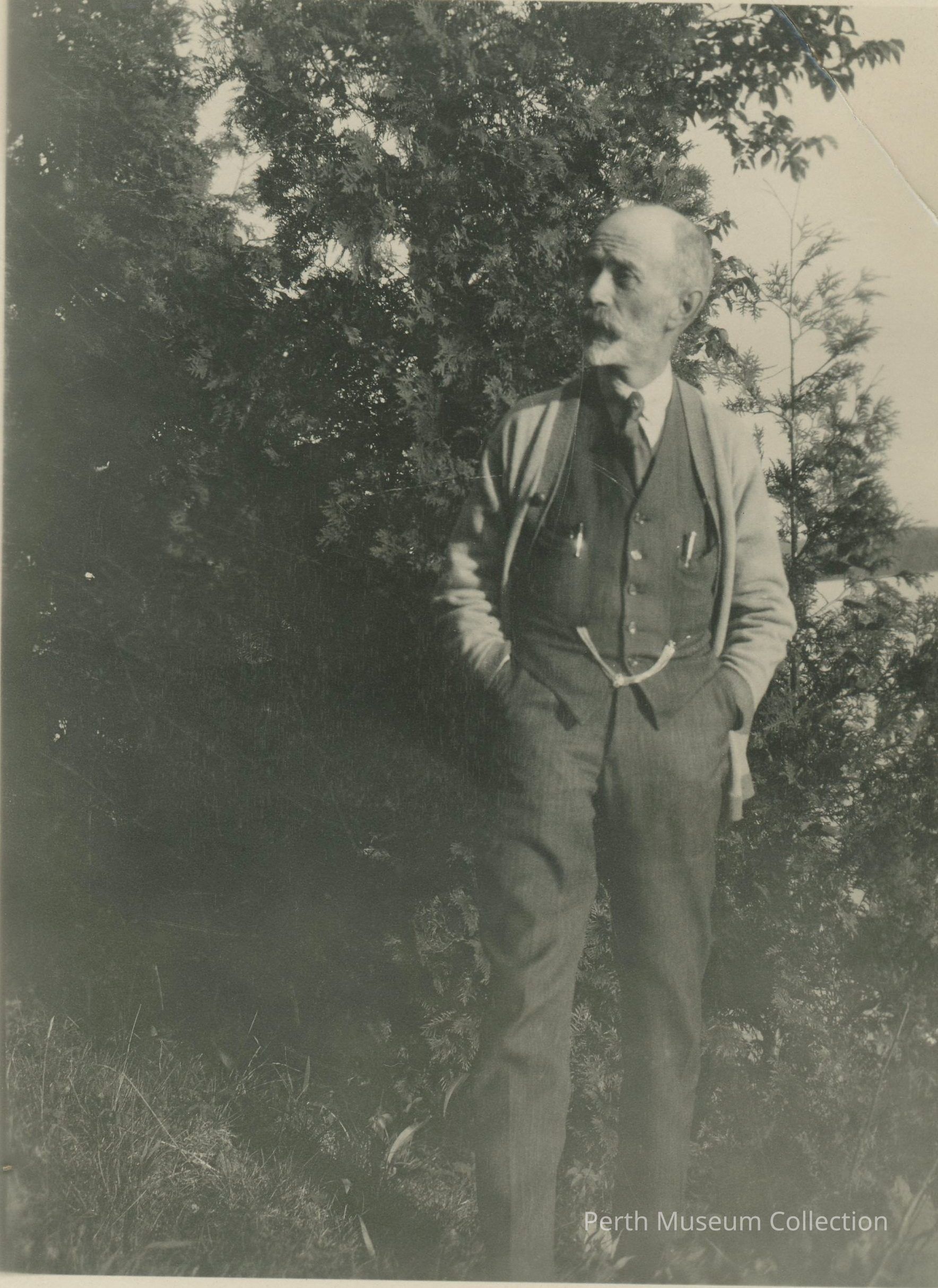 Archibald Campbell: The black-and-white photograph depicts an older man with a mustache standing outdoors, dressed in a three-piece suit with a pocket watch chain, and an open cardigan, hands casually in his pockets, gazing thoughtfully to the side, surrounded by lush foliage from large trees, suggesting a serene and contemplative moment in nature, as sunlight filters through the leaves, casting dappled shadows on the ground, contributing to the tranquil and timeless ambiance of the scene, reflecting a sense of calm and introspection.