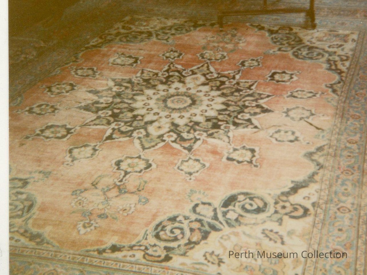 Rug: The image depicts an ornate rug with a large, intricate central medallion design featuring symmetrical floral patterns and complex geometric shapes, primarily in shades of dark blue, black, and cream, set against a muted red background. The border of the rug is adorned with smaller motifs and a repeating pattern, contrasting the more elaborate centerpiece. The wear and slight discoloration of the rug suggest age, adding to the vintage aesthetic. Dim lighting enhances the warm tones, contributing to the overall antique feel of the scene.