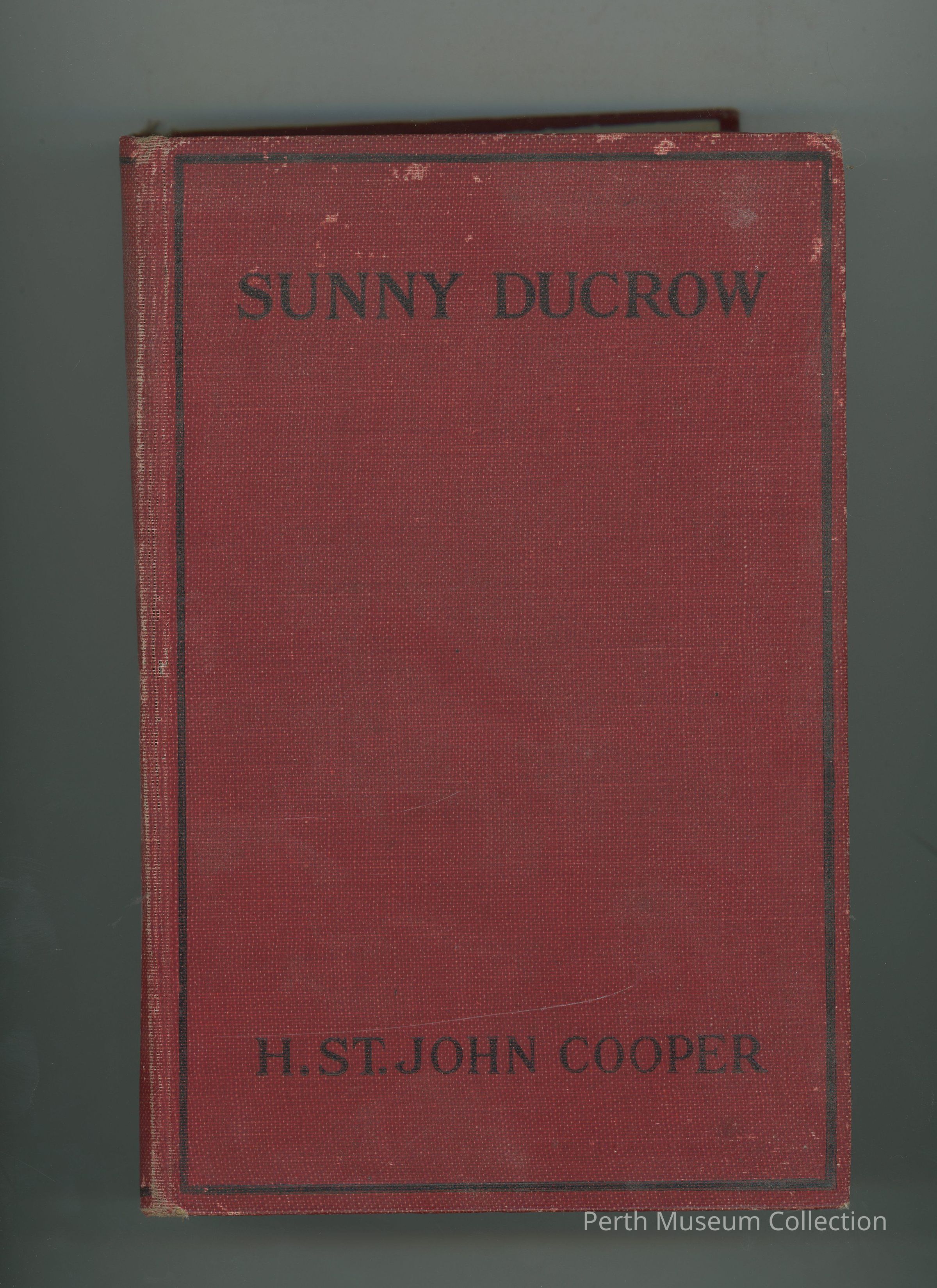 Cover: The image displays the front cover of a hardcover book titled "Sunny Ducrow," authored by H. St. John Cooper. The cover is a deep, solid red color, featuring two black rectangular borders that frame its edges. The title is prominently printed near the top in bold, black uppercase letters, while the author's name is similarly styled at the bottom of the cover. The book appears to be used, with visible wear along the spine and edges, indicative of a well-loved, vintage copy.