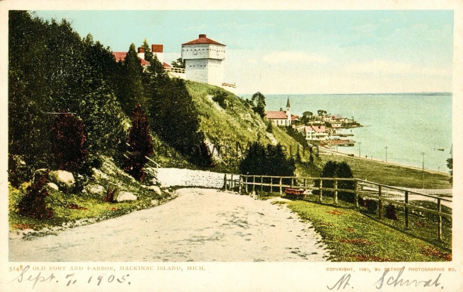 Postcard, Old Ford and Harbor, Mackinac Island, Mich: The image is an old postcard depicting a scenic view of Mackinac Island, Michigan, dated September 7, 1905 featuring a winding path flanked by greenery, leading up to a historic fort situated atop a hill, with its distinct white structure and red roofs clearly visible; below the hill, there's a cluster of buildings and a church near the coastline, creating a picturesque backdrop against the expanse of a tranquil, blue lake extending to the horizon. Copyright 1899 by Detroit Photographic Co.