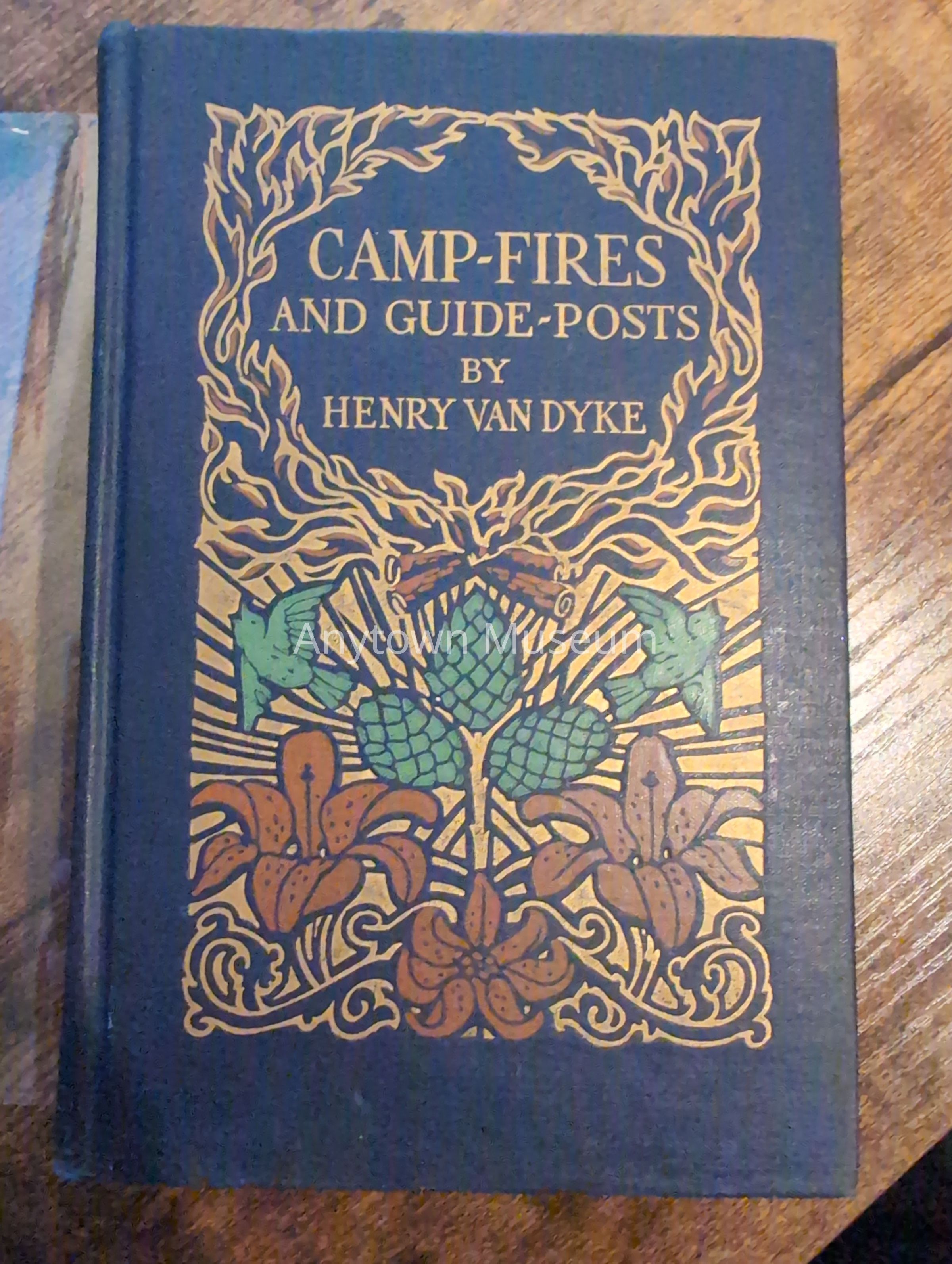 Front cover of a cloth-bound book with gilt lettering and decorative botanical illustration. The cover features an ornamental frame of stylized foliage surrounding the title, author name, and central motif of hops and floral elements rendered in green, orange, and gilt.: This is a hardcover book bound in blue cloth with gilt decoration on the front board. The cover displays the title "Camp-Fires and Guide-Posts" and author attribution "by Henry van Dyke" in gilt lettering arranged within an ornamental rectangular frame. The frame consists of stylized, interlocking organic forms suggesting flames or foliated vine work, rendered in gilt against the blue ground. Within the frame, a botanical composition occupies the central lower portion, featuring two green hop cones with characteristic scaled forms and broad green leaves flanking a bundle of overlapping brown flowers, possibly lilies or similar decorative blooms, positioned symmetrically. The composition extends downward to ornamental scrollwork in gilt and rust tones at the base. The spine is visible as a raised ridge along the left edge, indicating a case-bound construction typical of early-to-mid 20th-century publishers' bindings. The overall design is consistent with Arts and Crafts and Art Nouveau aesthetic traditions, emphasizing nature-inspired ornamentation and refined hand-lettering. The book rests on a wooden surface, and minor wear is visible on the cloth and edges, consistent with age and use. The gilt decoration shows selective fading and wear appropriate to an antiquarian volume.