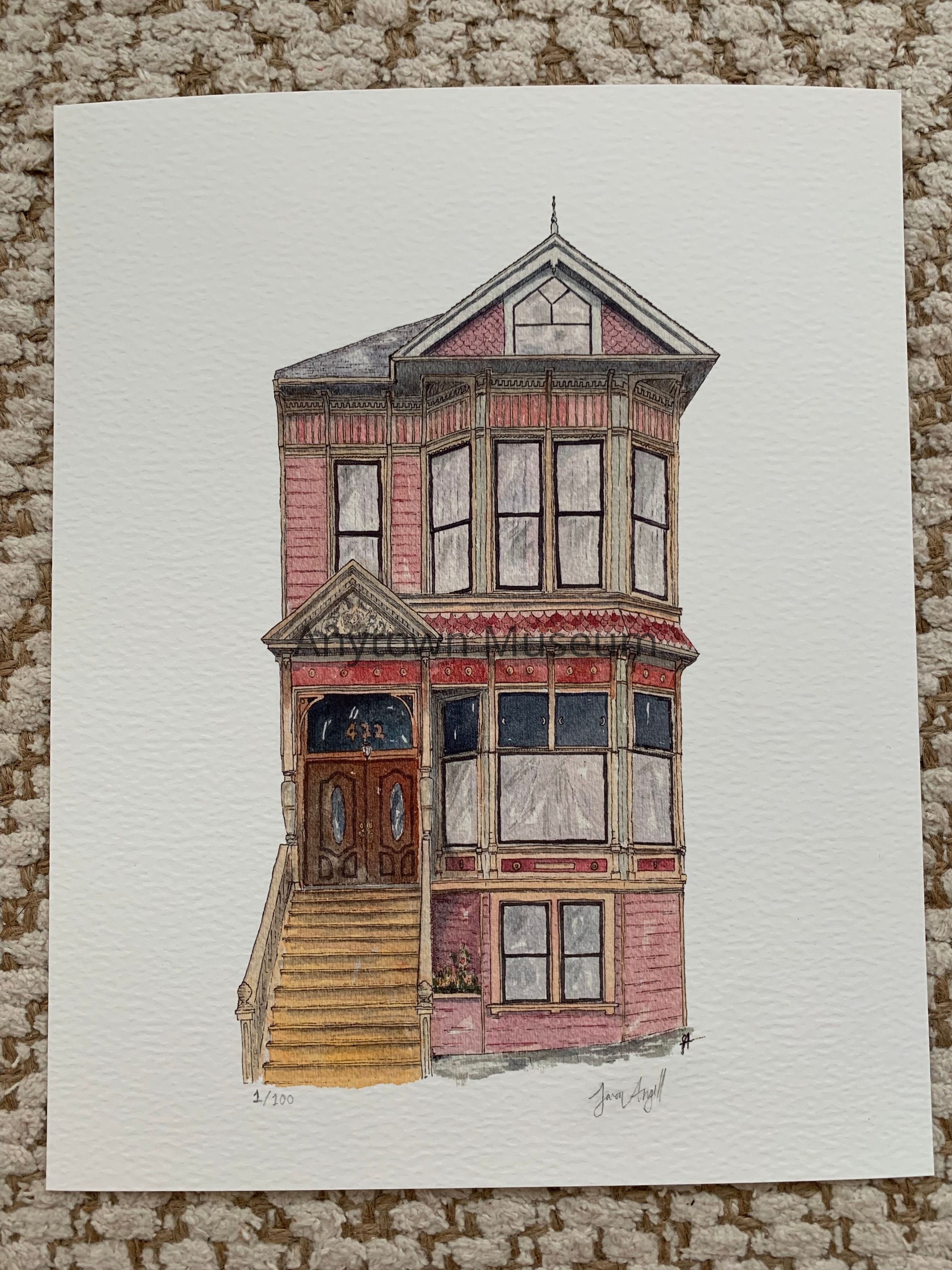 A precisely rendered architectural illustration depicts a narrow pink Victorian townhouse with ornamental details. The front-facing view shows a tiered structure with a peaked roofline, bay window, recessed storefront entrance with brown paneled doors, and large window panes across multiple levels. The print is mounted on white paper against a textured beige background.: This is a digital print reproduction of a watercolor architectural illustration depicting a three-story Victorian-era residential or mixed-use building. The structure features a distinctive narrow facade finished in pink brick or painted surface with decorative trim throughout. The composition is organized vertically, emphasizing the building's height. The lowest level contains a recessed storefront with dark double doors ornamented with inlaid ovals, flanked by multipane windows and topped with decorative molding and signage band. A wooden staircase with multiple steps provides access to this entrance. The middle story features a prominent bay window with multiple large panes arranged in a grid pattern, set within a projecting frame with decorative cornice detailing in dark tones and ornamental brackets. The upper story mirrors similar window proportions but in simpler form, with a peaked gable roof crowned by a spire finial. Throughout the facade, decorative dentil moldings, applied ornament, and painted details suggest Victorian-era commercial or residential architecture, likely from the late 19th or early 20th century. The artist has employed watercolor washes and ink line work to articulate architectural elements, materials, and dimensional relief. A printed notation "1/100" at lower left indicates this is a numbered reproduction from an edition. An artist's signature appears at lower right. The print is rendered on white paper with visible paper texture and is displayed against a textured neutral background.