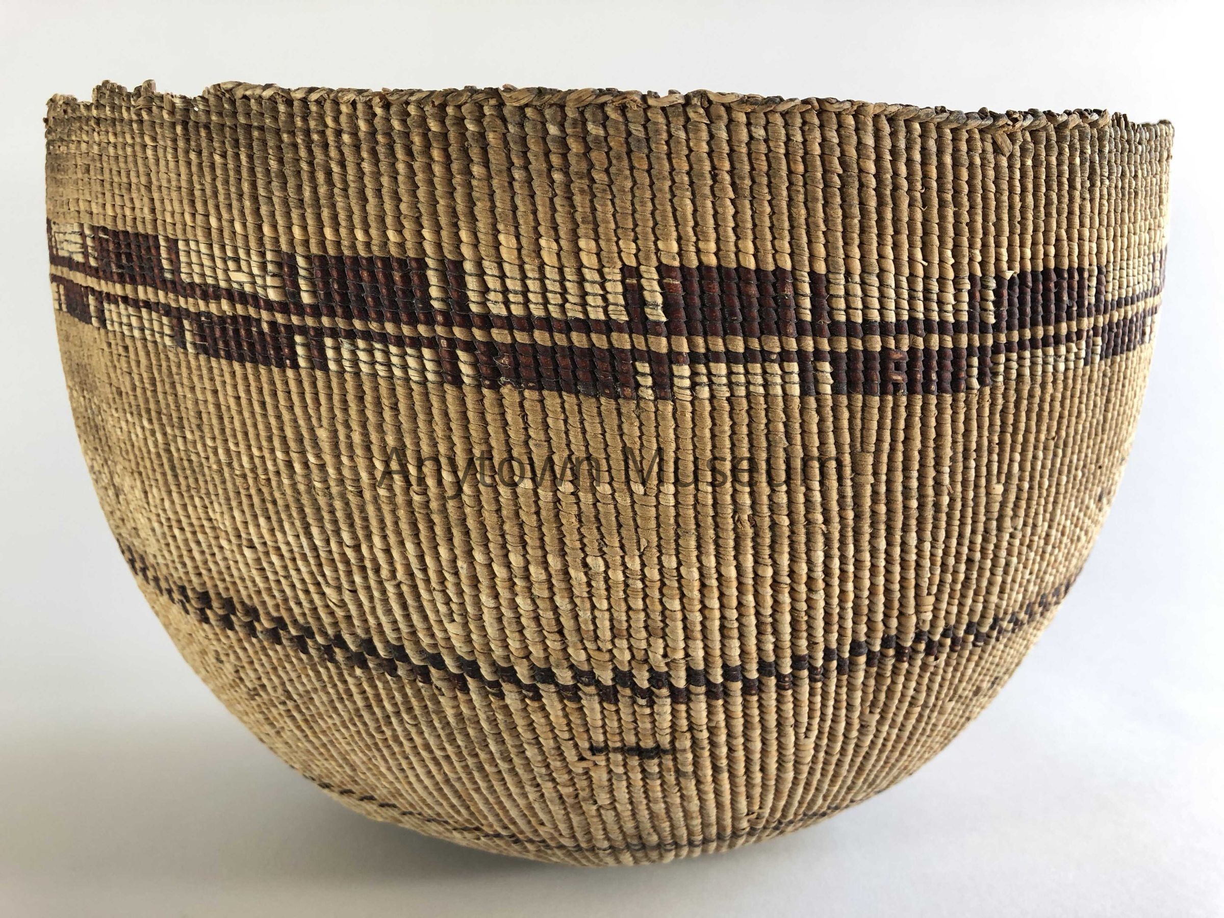 A tightly woven basketry vessel with a rounded, deep bowl form displays rectangular and square patterns in dark and light twining across its surface. The basket is presented in three-quarter view against a neutral background, highlighting its dense weave and subtle geometric decoration.: This cooking basket exemplifies Pomo basketweaving traditions of Northern California. The object is a hemispherical or bowl-shaped vessel constructed using the twining technique, a method in which horizontal weft elements are twisted around vertical warp strands. The basket exhibits a dense, regular weave characteristic of utility cooking baskets designed to hold liquids and withstand direct heat. The exterior surface is decorated with bands of rectangular and square patterns created through the use of contrasting materials—likely natural willow or sedge in light tones combined with darker-colored plant fibers, possibly redbud, to create the geometric designs that encircle the vessel. The patterns are organized in horizontal registers and do not feature the zigzag or diamond motifs sometimes found in Pomo basketry; instead, the design is based on rectilinear forms aligned with the structural grid of the twining itself. The rim appears finished with a binding or wrapped edge typical of functional vessels. The overall form suggests the basket was intended for cooking acorns, either for leaching, parboiling, or rendering the nuts for food preparation. The construction is executed with considerable technical skill, with uniform spacing and tension throughout. The object shows some age-related darkening and surface wear consistent with use and age, particularly visible in areas of the darker patterning.