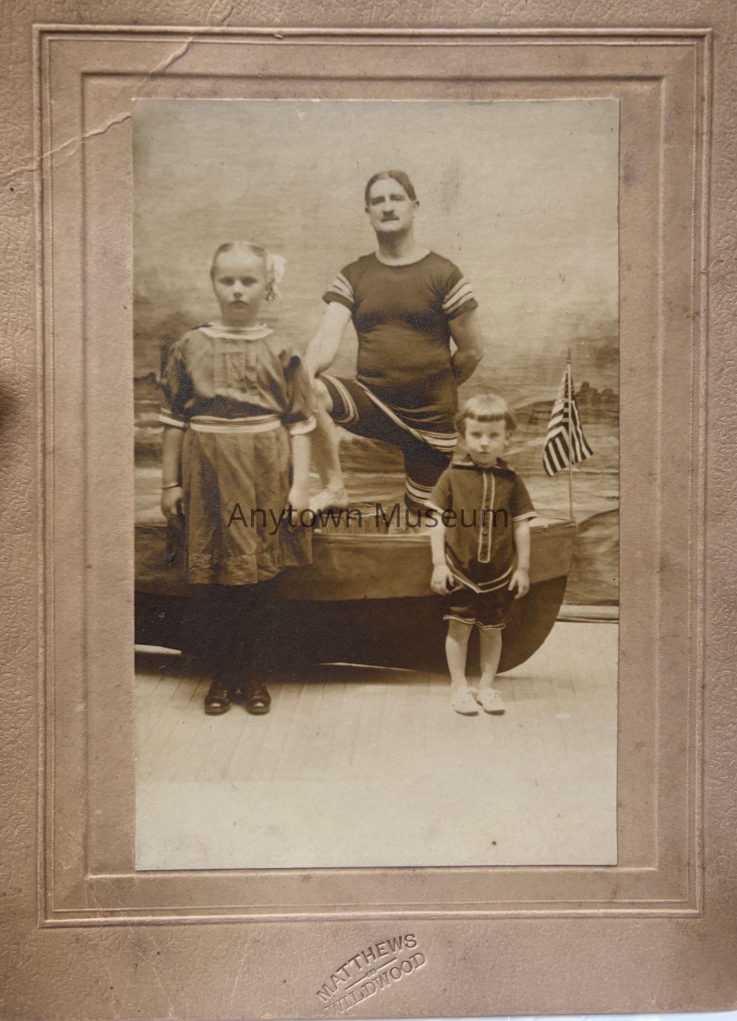 The vintage photograph features a mustachioed man in a striped bathing suit standing confidently on one leg, beside two children wearing period dresses and boots, with a painted seascape backdrop including a small boat and American flag, all framed with a textured border and labeled "Matthews Wildwood," evoking a nostalgic early 20th-century seaside vibe, capturing an intriguing glimpse into past fashion and family moments shared by these three individuals at a likely seaside location, portraying a sense of tranquility and timelessness.
