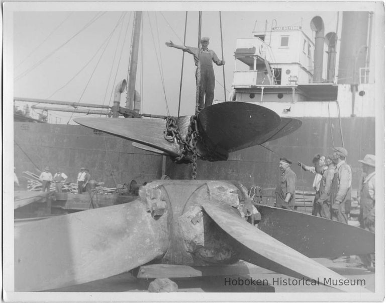 B+W photo of a worker standing on horizontal 4-blade propeller (a new or refurbished one) being moved by a crane at the Bethlehem Steel Shipyard, Hoboken, no date, ca. 1940. picture number 1