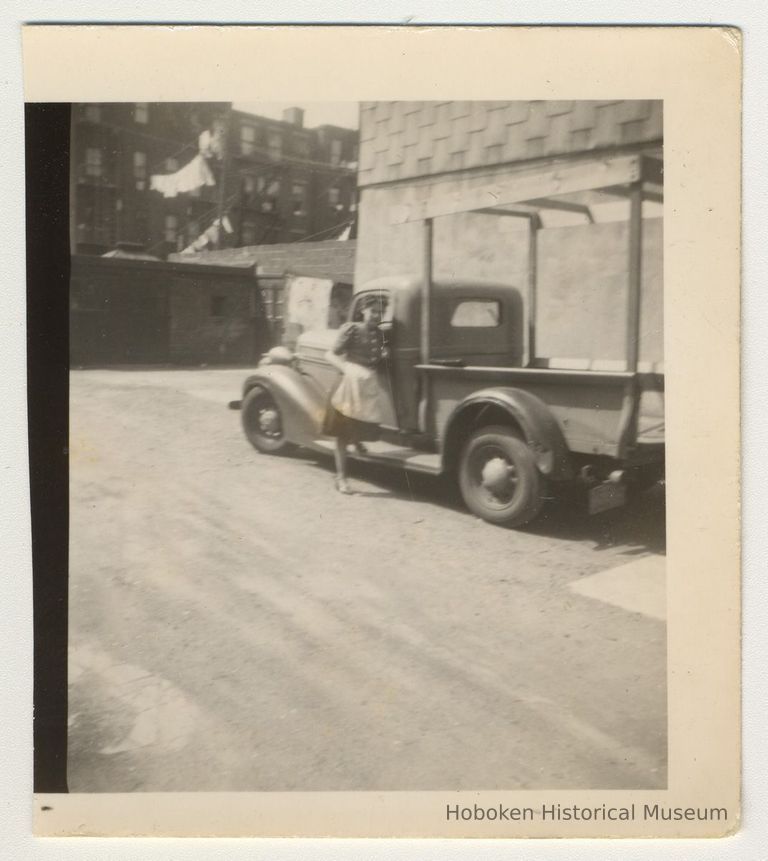 Digital image of b+w photo of Ann D'Atilla standing next to a Lemon Ice & Yum Yum truck in the backyard, 416 Clinton St., Hoboken, n.d., ca. 1950s. picture number 1