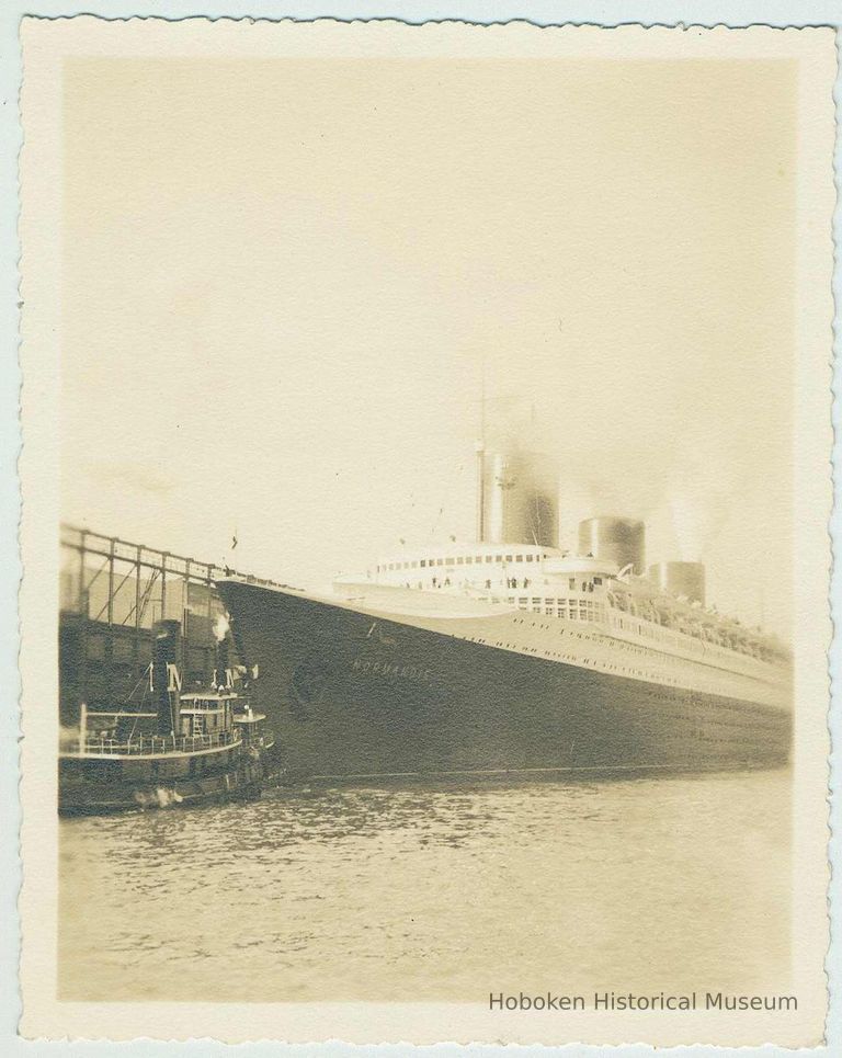 B+W photo of the ocean liner S.S. Normandie at a pier under steam with tugboat at bow. N.d., ca. 1935-1939. picture number 1