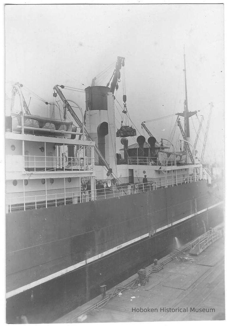 B+W photo of a view amidships of an unknown ship at berth; cargo is being hoisted at center. picture number 1