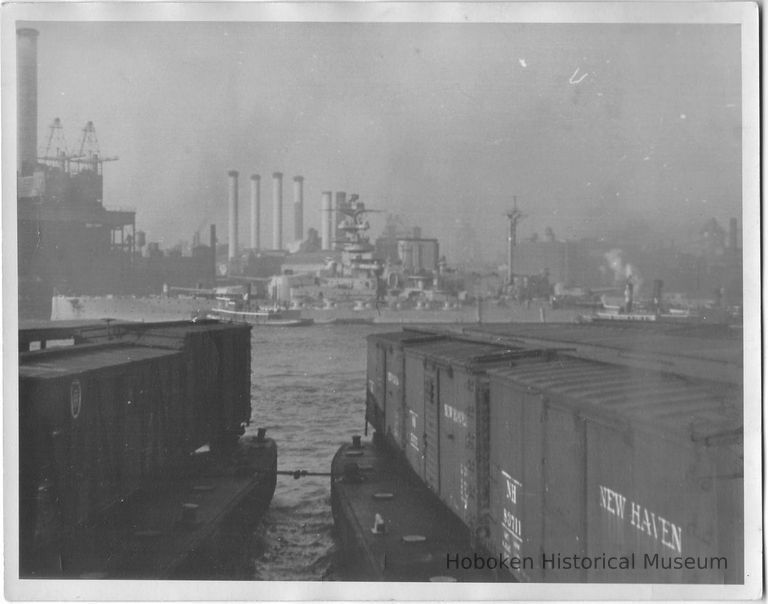 B+W photo of a warship with tugboats in the East River of New York City; car floats in foreground, N.Y., no date, ca. 1940. picture number 1