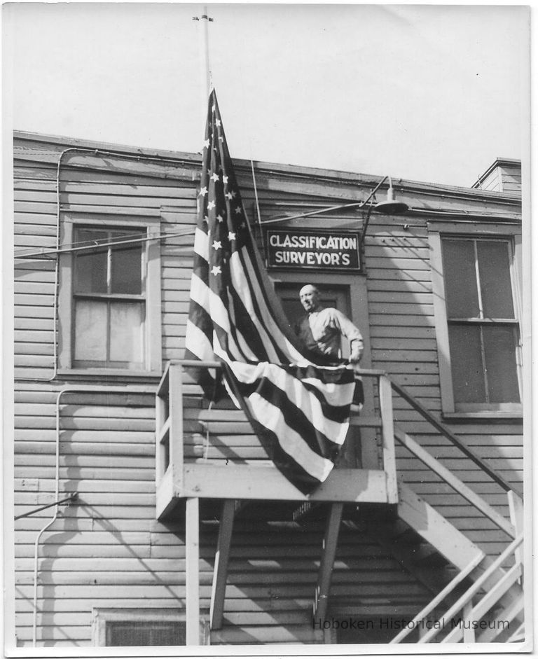 B+W photo of the Classification Surveyor's office exterior with a man standing next to a flag, Hoboken, no date, ca. 1940. picture number 1