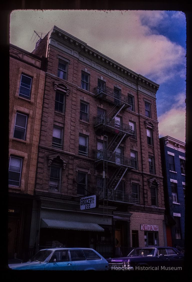 Color slide of eye-level view of brick façade, cornice, pediments and fire escape at 202-204 Hudson between 2nd & 3rd showing a grocery store and the India Bazaar storefronts picture number 1