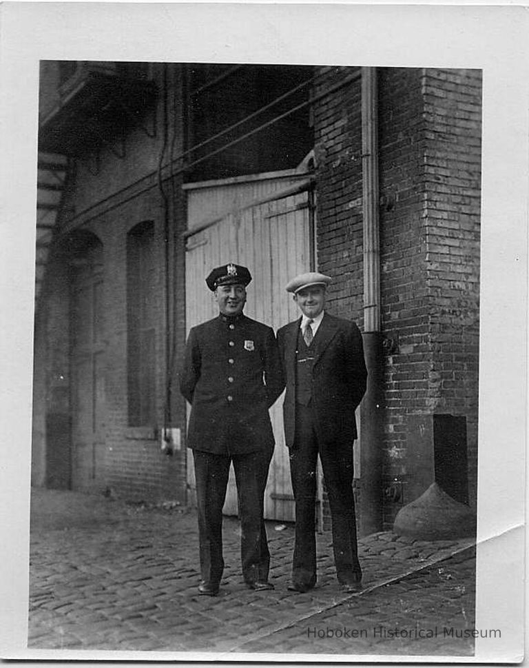 B+W photo of a policeman and man with cap posing in front of brick building, Hoboken shipyard, no date, ca. 1940. picture number 1