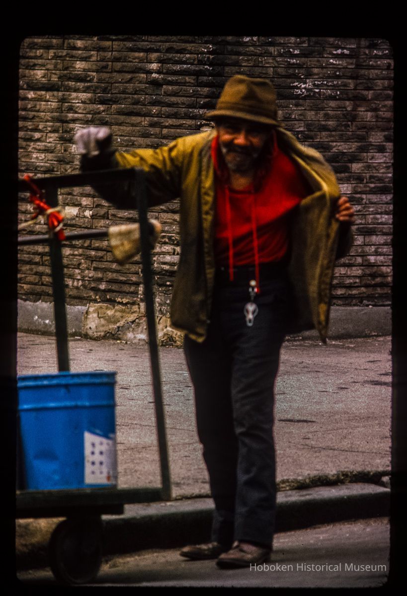 Color slide of eye-level view of a man in a red shirt, yellow jacket, black pants and a brown hat pushing a cart on Park and 1st picture number 1