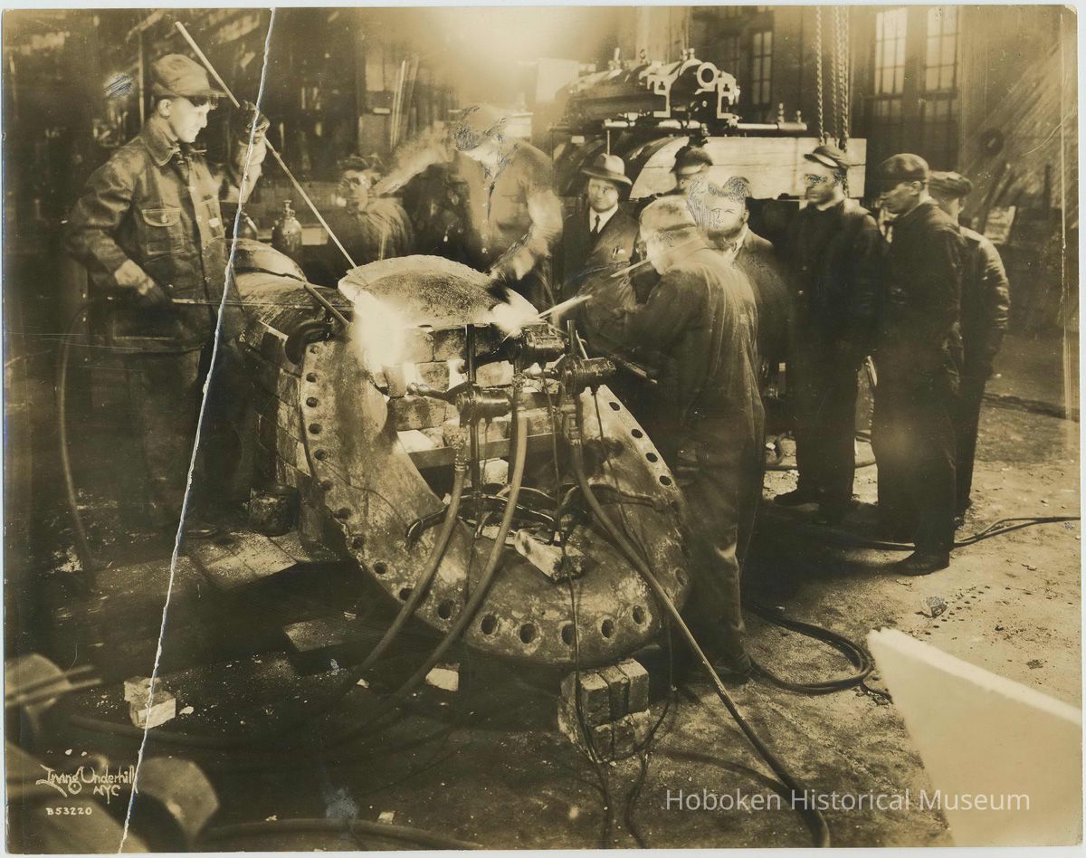Sepia-tone photo of workers using torches on a large ship fitting in a shop at the W. & A. Fletcher shipyard, Hoboken, no date, ca. 1925. picture number 1