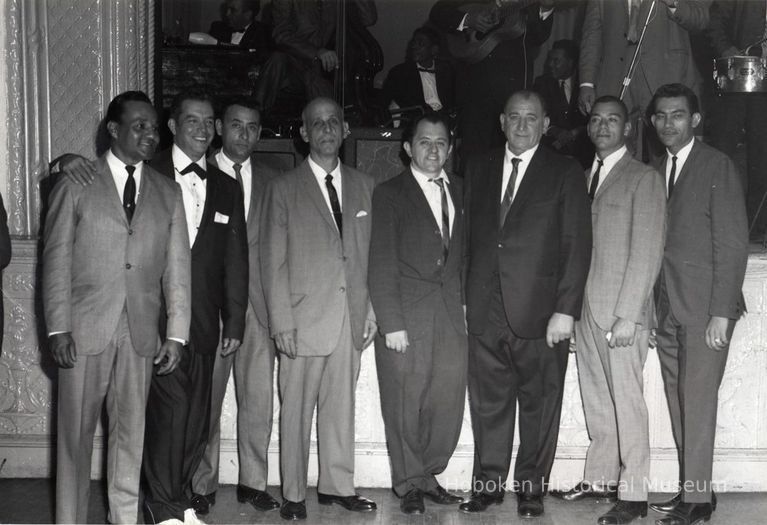 B+W group photo of eight men posed in front of a stage, (Hoboken), no date, ca. 1960-1970. picture number 1