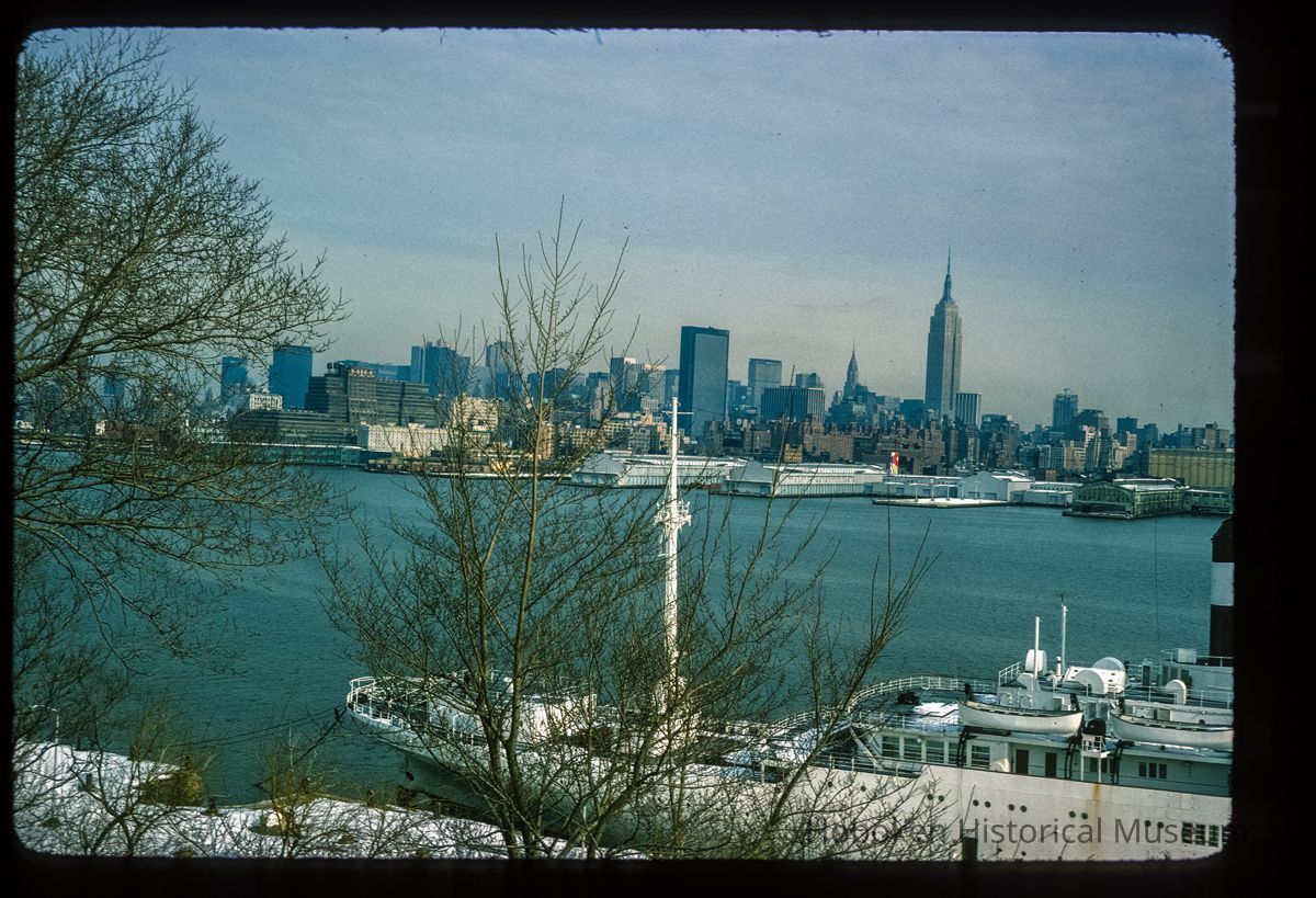 Color slide of eye-level view from the Stevens Institute campus of the SS Stevens docked at Castle Point with the New York City skyline including the Empire State Building across the Hudson River picture number 1