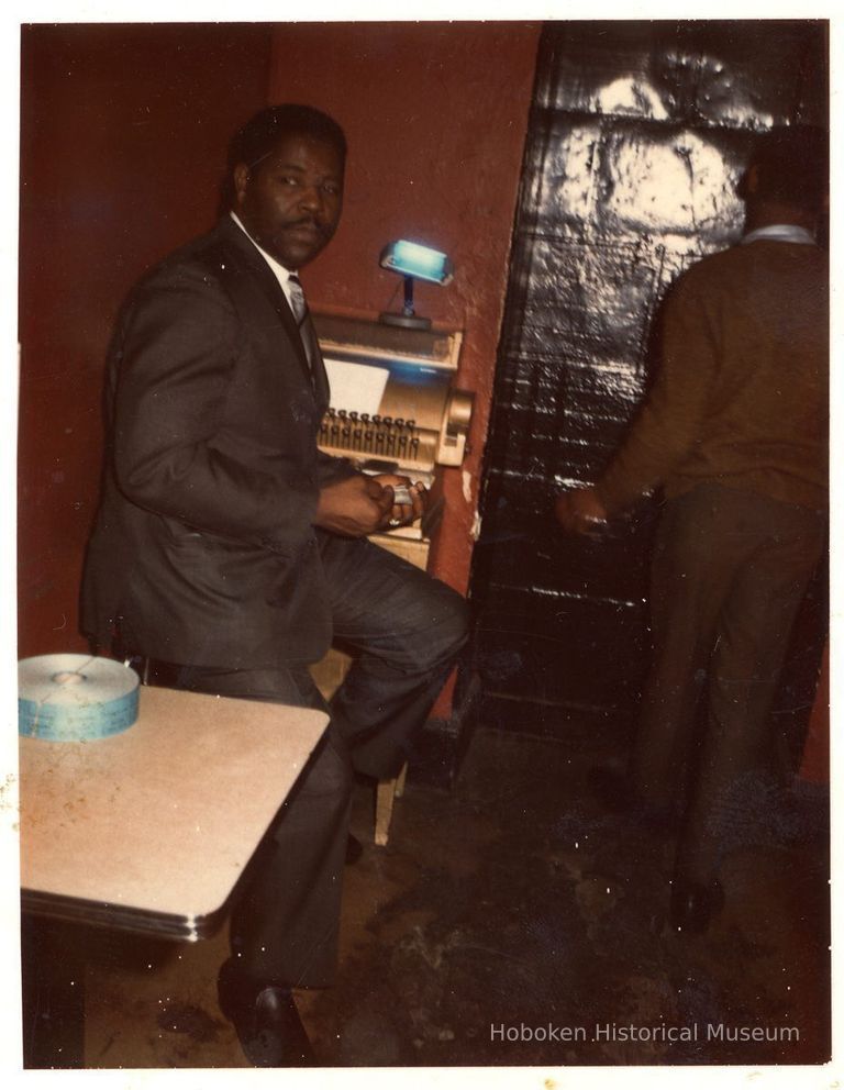 Digital copy of color photo of Charles McNeil seated at the a cash register, Zanzibar Club, Hoboken, circa late 1960's. picture number 1