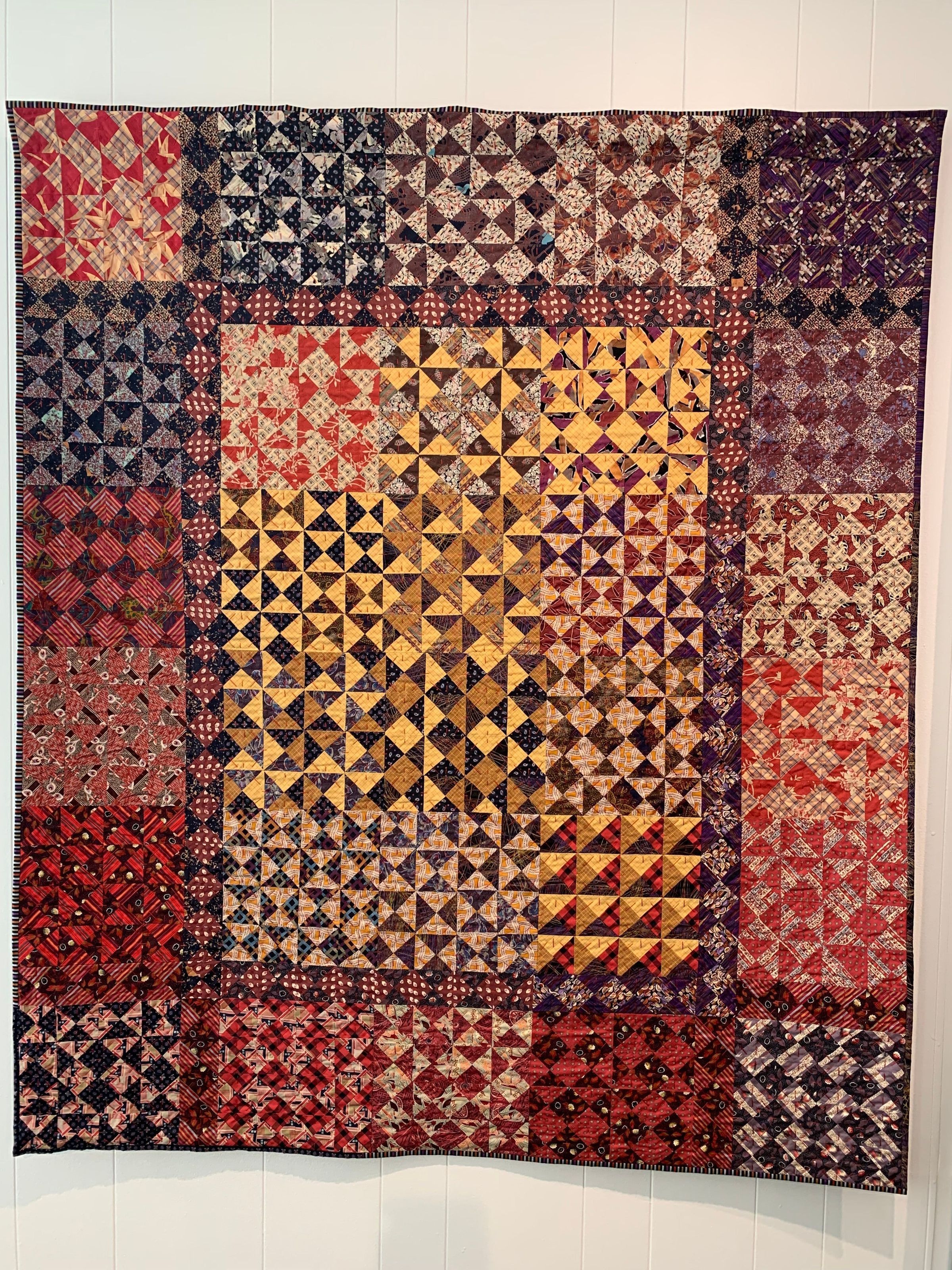 The quilt on display is a vibrant patchwork of various geometric patterns and colors, predominantly featuring shades of red, yellow, blue, and brown, arranged in a symmetrical grid. It showcases a mixture of intricate hourglass designs, including triangles and squares, with each section presenting a unique combination of motifs and colors. The overall design creates a dynamic visual effect, reflecting the traditional craftsmanship and artistic expression involved in its creation. The quilt is bordered in a darker hue, providing a frame that complements the diversity of patterns within.