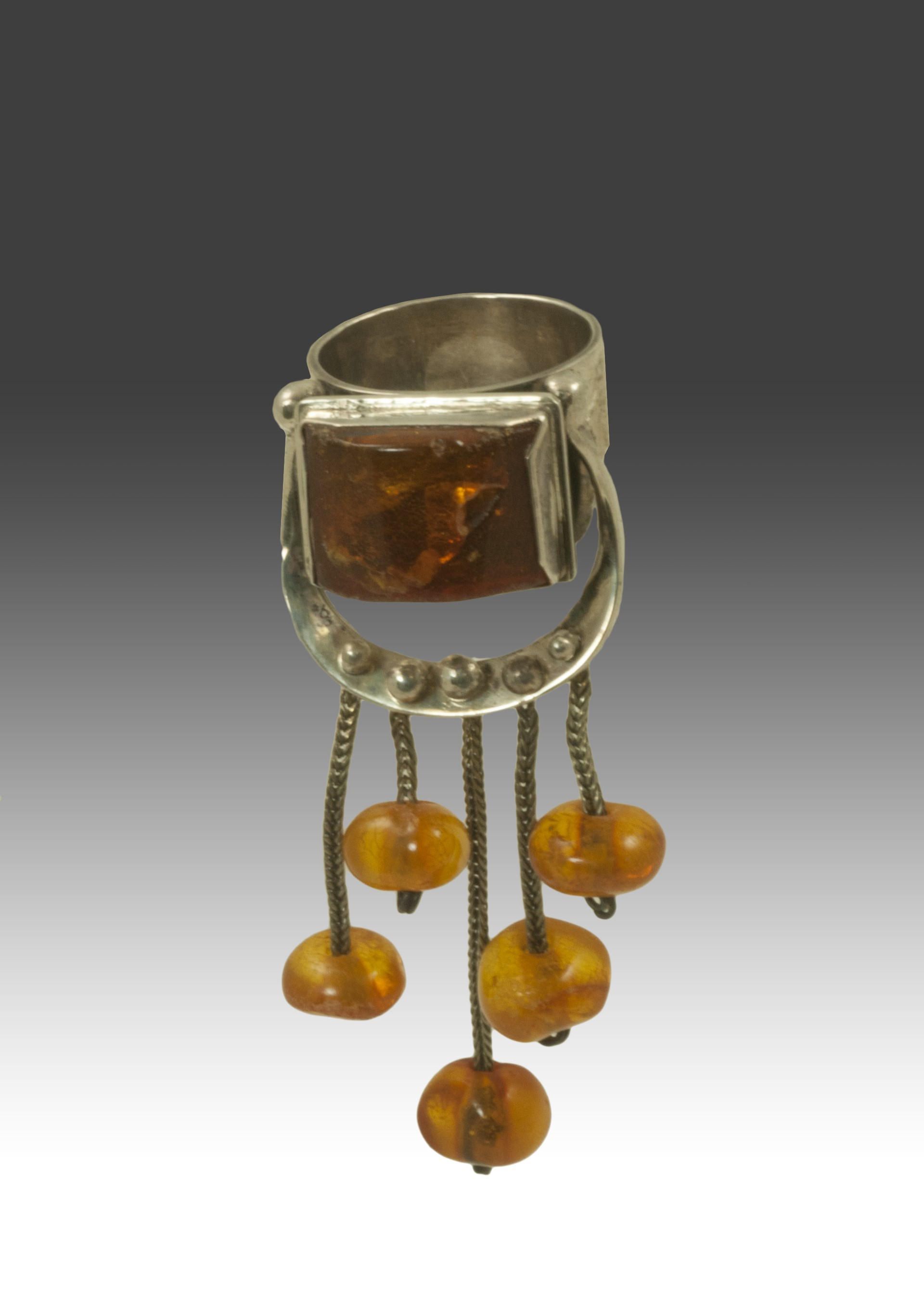 The image shows a unique piece of jewelry—a ring with an intricate design featuring a large, raised silver setting that encases an amber stone. Dangling from the setting, there are small chains attached to which more amber beads are affixed, creating a chandelier effect. The silver setting is adorned with decorative elements, giving the ring an ornamental, vintage appearance.