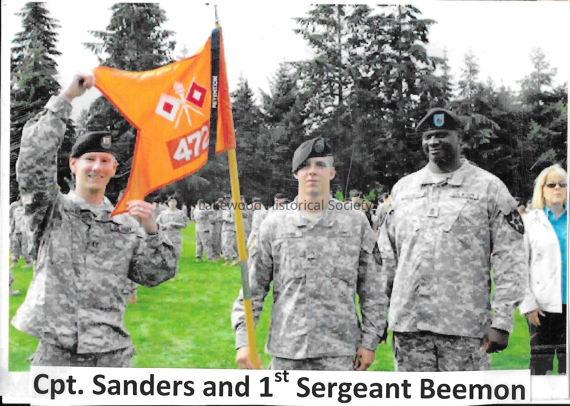 Three men in military field uniforms holding the flag for the 472nd: 2024.002.027 - Color photograph of three men standing in field with trees behind them. The men are Cpt. Sanders (left), 1st Srgt. Beemon (right) and a third unidentified man (middle). The man in the middle hols a pole with the 472nd's flag attached. Sanders is holding the flag out for the camera. A woman in a blue shirt and white jacket can be seen behind Beemon.