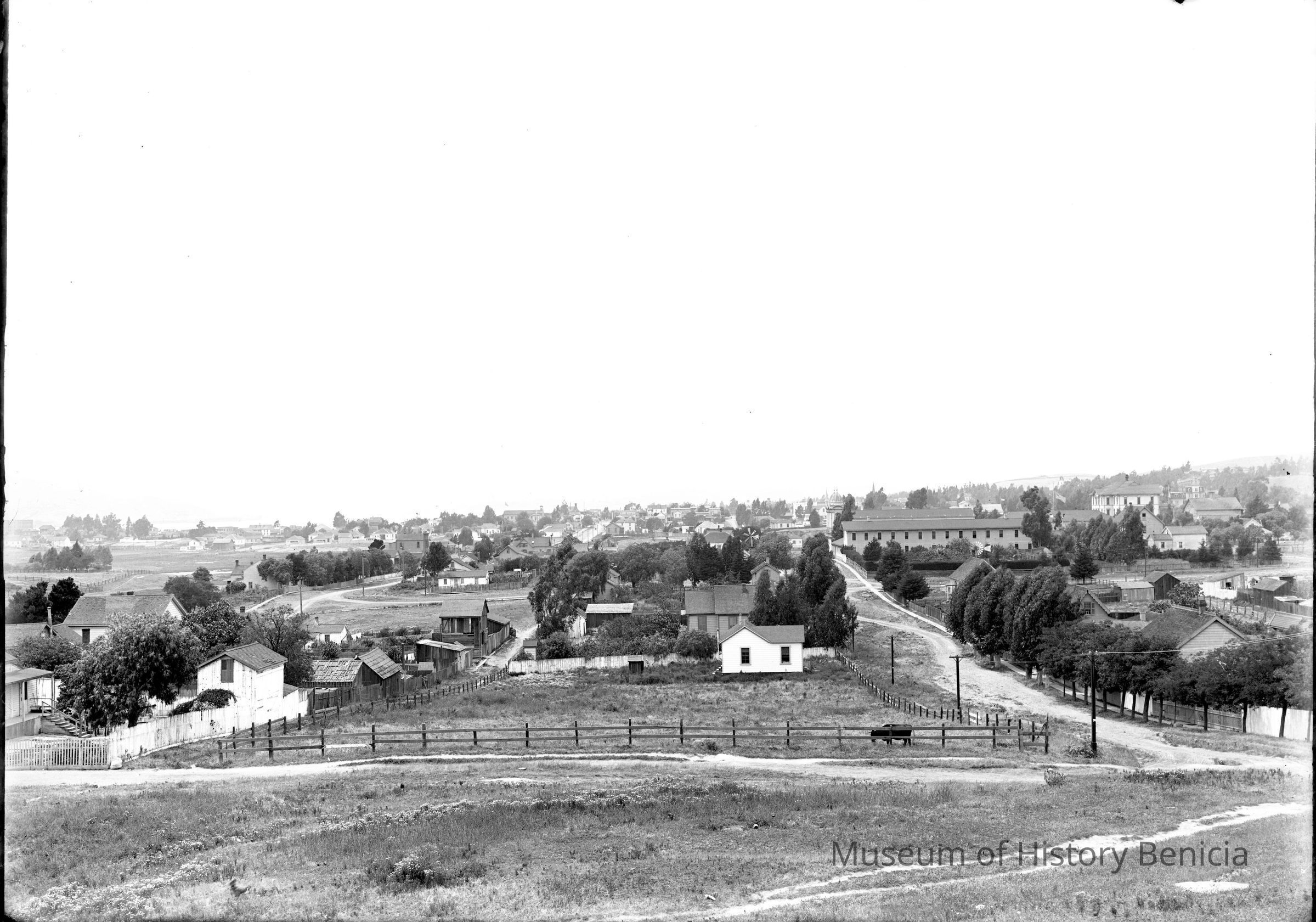 The image depicts a panoramic view of Benicia, California, around 1910, showcasing a quaint and serene small town scene. In the foreground, expansive grass fields are dotted with small wooden houses and fenced-in gardens, illustrating the rural charm and simplicity of early 20th-century life. Winding dirt roads lead through the landscape, connecting clusters of modest homes and more substantial buildings, which likely serve as community centers or commercial establishments. Rows of trees line some of the streets, providing shade and structure to the area. The background reveals a sprawling townscape, with more residences interspersed among open spaces, suggesting a balance between development and natural surroundings. The gentle undulations of the land, along with the scattered trees, create a sense of tranquility and openness. The architectural style of the houses, characterized by simplistic designs and wooden construction, reflects the practical yet quaint aesthetics prevalent during this period in American small towns. The photograph captures a moment in time, conveying the peaceful yet industrious nature of early