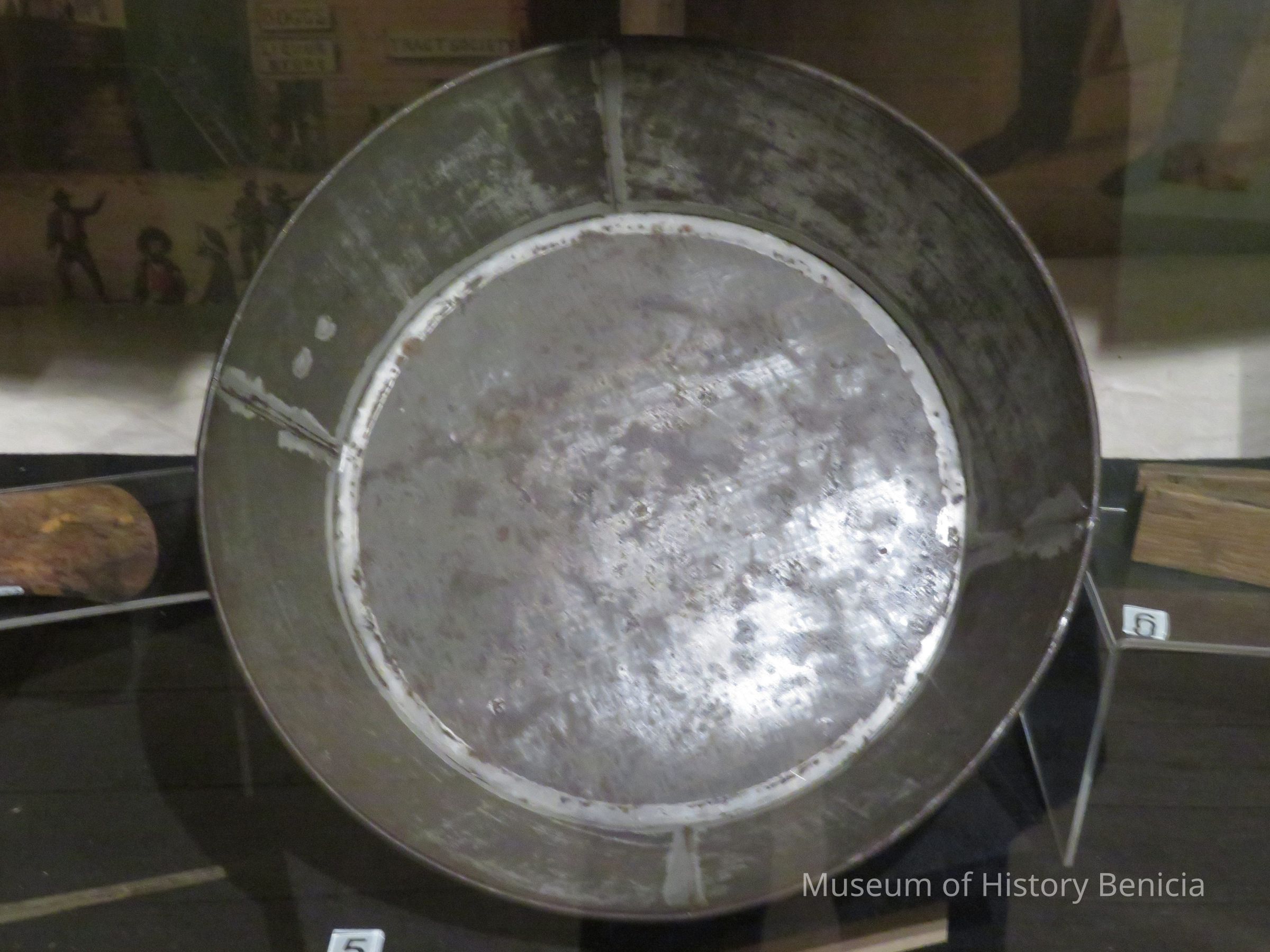 The image shows an old, metallic gold pan used historically for panning gold, featured in a museum display. The pan, which is wide and circular, appears well-worn, indicative of extensive use, with scratches and tarnished areas visible on its surface. It sits upright, supported by a stand, and is surrounded by additional exhibits, hinting at its historical significance during gold rush periods. Behind the pan, a backdrop with vintage illustrations or photographs provides context to its usage, possibly depicting scenes of prospectors at work in the 19th century. This setup highlights the importance of such tools.