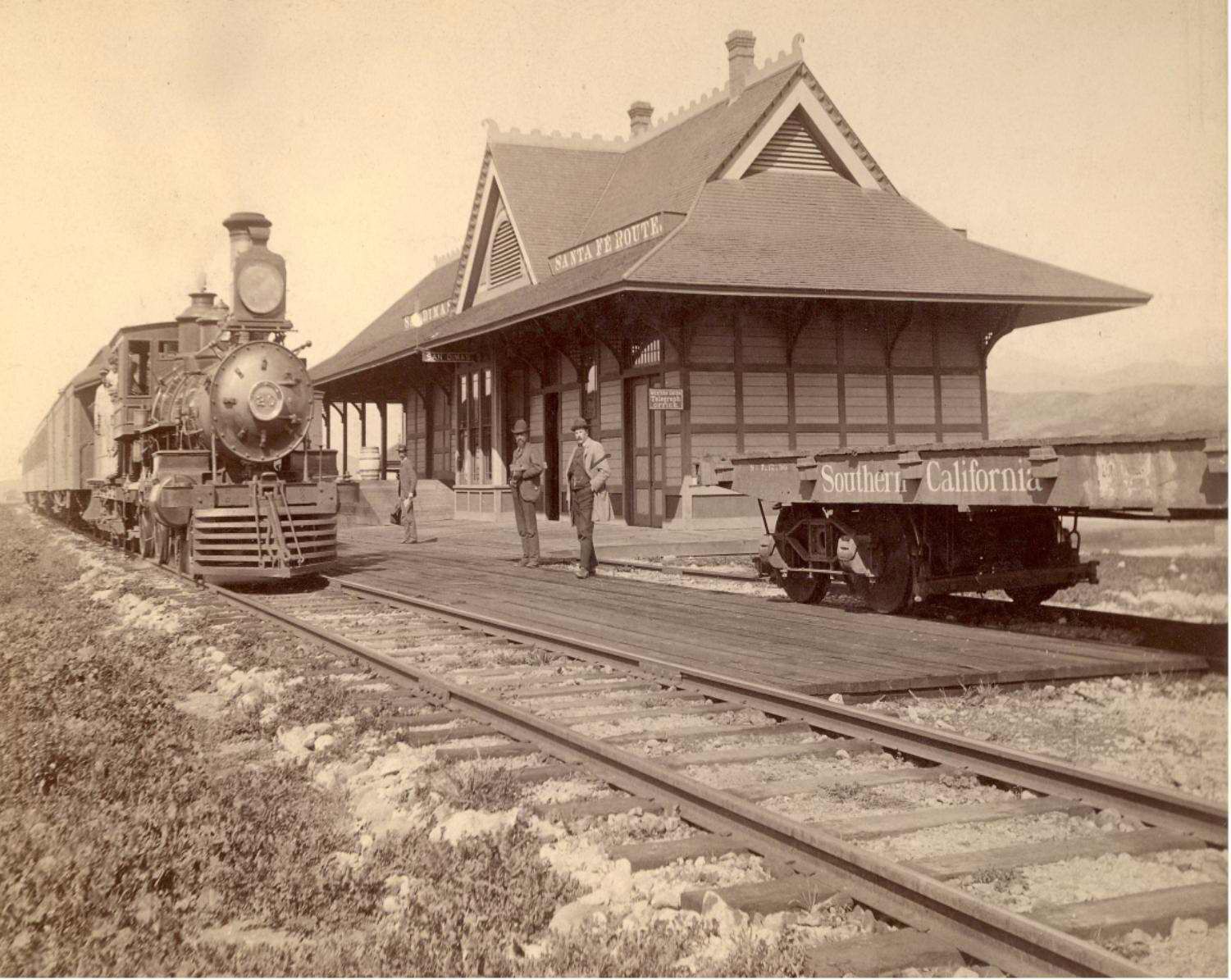 J-0070-0001: Santa Fe Railroad ran the first train thru San Dimas in May of 1887. The Station was completed in 1889. A freight car was used as a depot prior to this. Thomas E. Gore was first station agent seen in photo with mail bag. J. H. Judson coming home from CA to Ohio after overseeing the laying out of his orange grove at San Dimas CA was killed here by train.