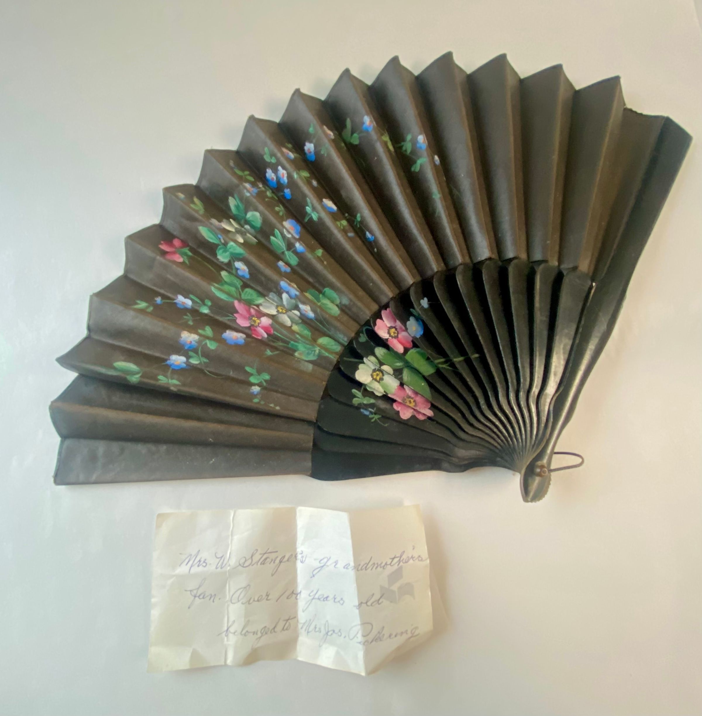 The image shows a beautifully crafted antique fan with intricate floral designs painted on its black face, featuring pink, blue, and green hues that add a touch of elegance and color. This hand fan, likely of significant age and historical value, is accompanied by a note written on a small piece of paper, providing context or provenance regarding the fan.