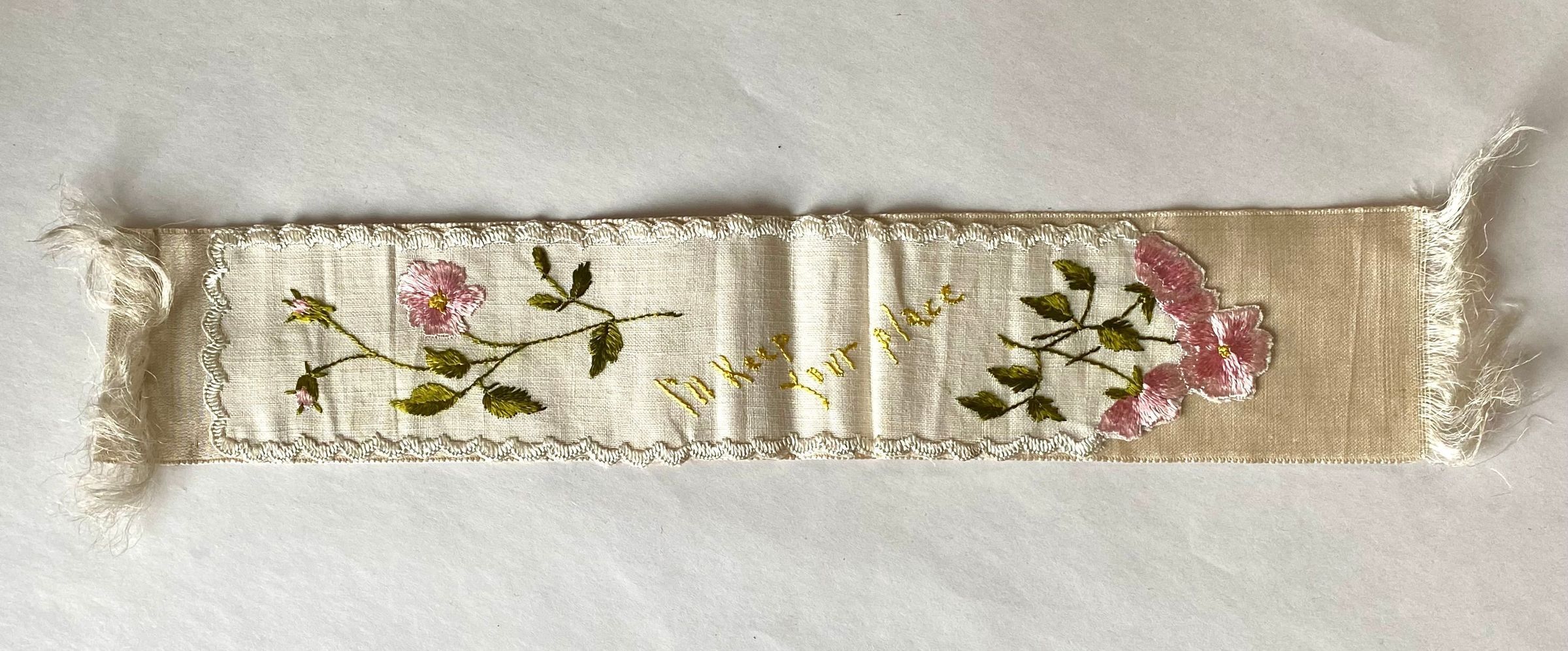 The image shows a delicate, vintage fabric bookmark featuring intricate embroidered pink flowers and green leaves on a light background, with fringed edges and the embroidered phrase "I'll keep your place" in yellow thread, creating a charming and elegant design that adds a touch of nostalgia and personalization to the simple piece.