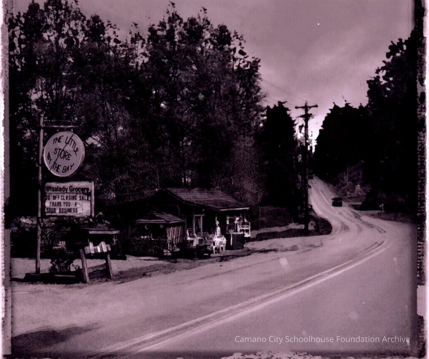 2024.003.I.002 The Utsalady Grocery was located at the corner of North Camano and Essex streets, facing North Camano.: A black and white photograph of a small brown wood store next to a curving road with a small parking lot, mailboxes, and a round sign that reads "The Little Store By The Bay," as well as a lettering sign below which is titled Utsalady Grocery and says "30% off closing sale, thank you for your business."
