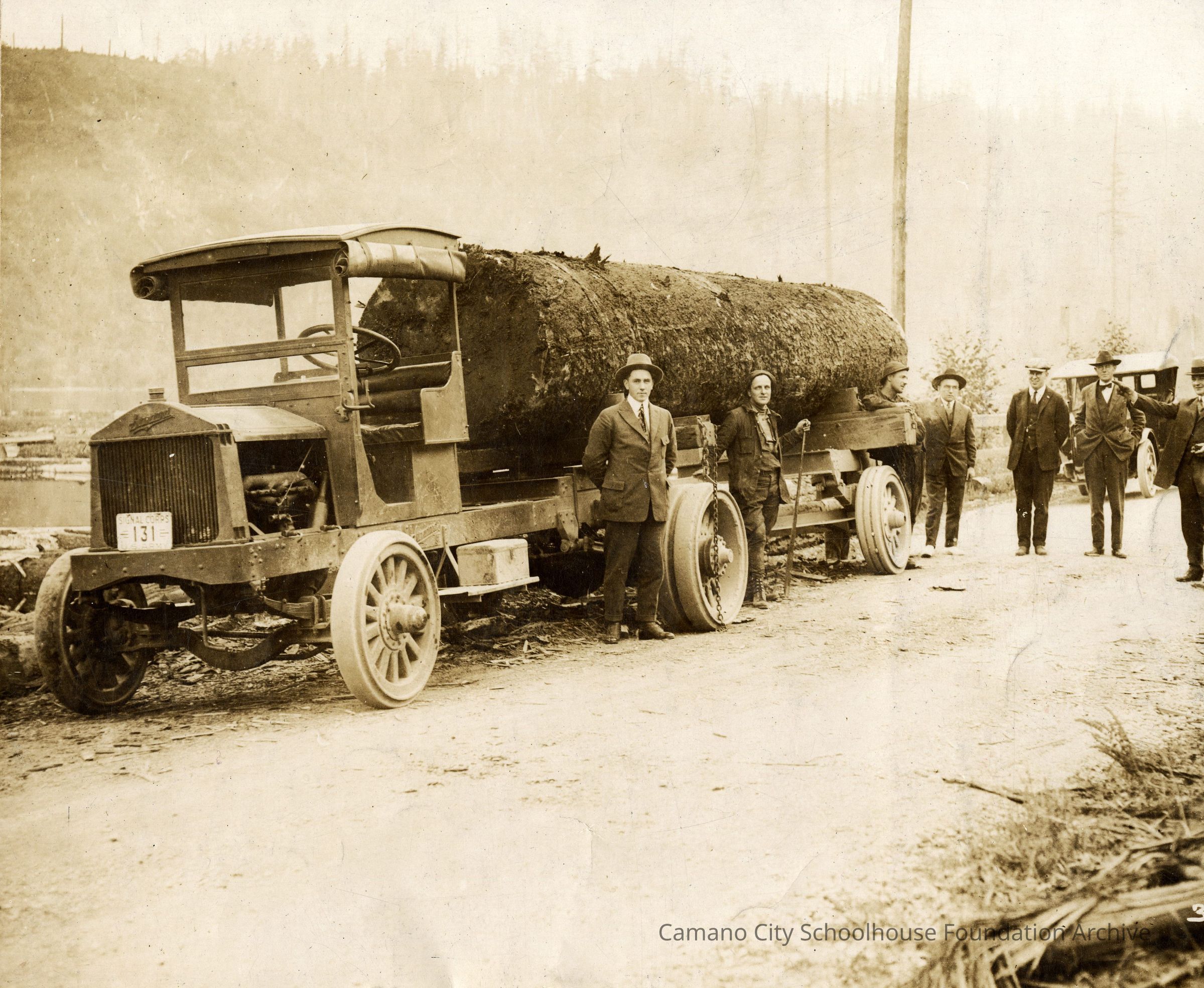 Six men stand around and behind an old-fashioned truck with a large (approximately 20 ft long by 5 ft across) log on the truck bed, ready to send off-island.: Six men stand in frame, most in suits with their hands behind their backs. They are spread around the right side of the photograph, and the left is dominated by an old-fashioned cargo truck with a large (approximately 20 ft long by 5 ft across) log on the truck bed. 2023.001.I.B2.a012