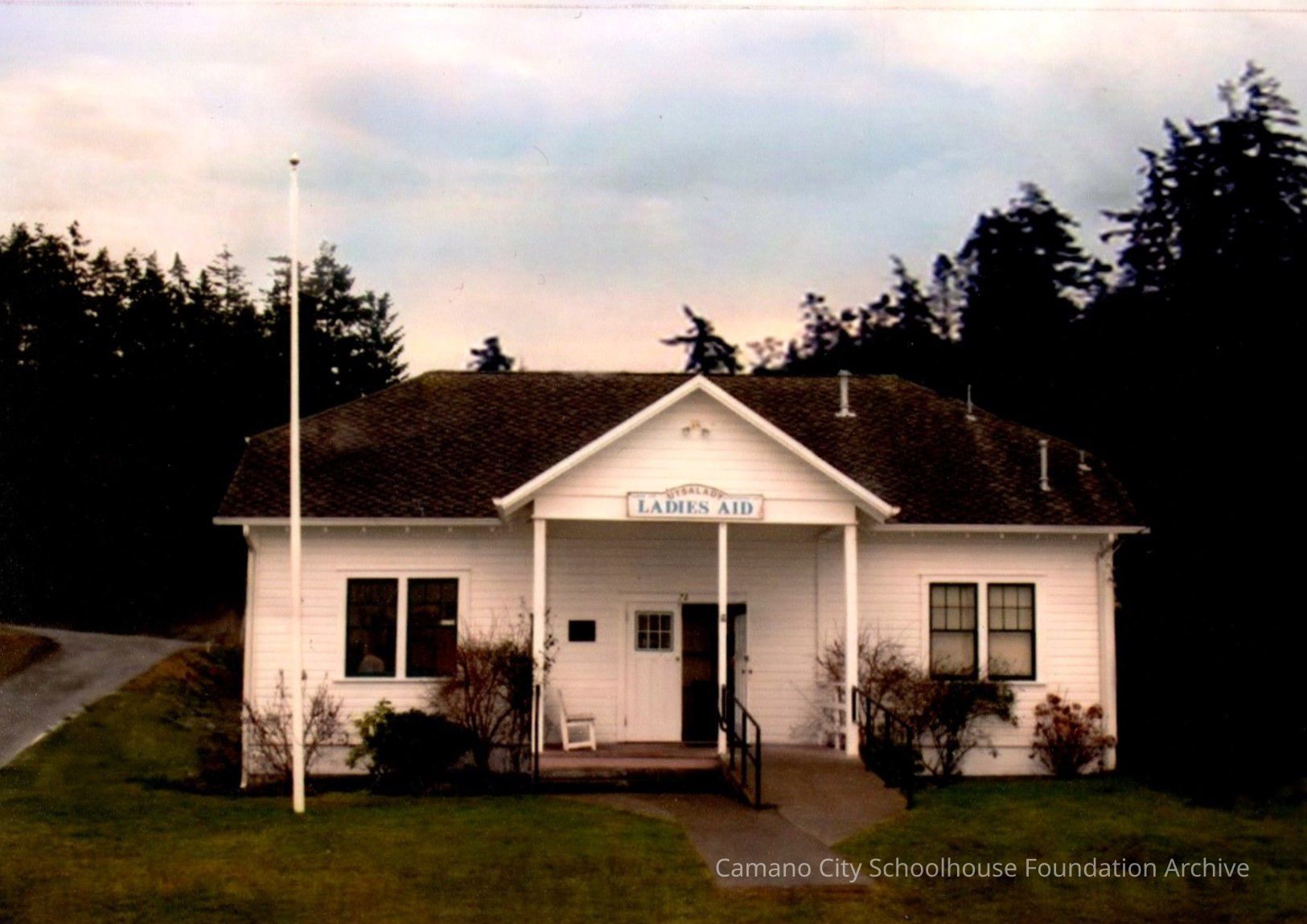 The Utsalady Ladies Aid building, which stands a the corner of Utsalady Road and Ekle Street, facing the water.: A white one-story wood building with a sign at the front roof peak reading Utsalady Ladies Aid, a ramp and steps to the door, and a flagpole in the grass yard. A gravel road is visible to the viewer's left and tall evergreen trees stand behind the building.