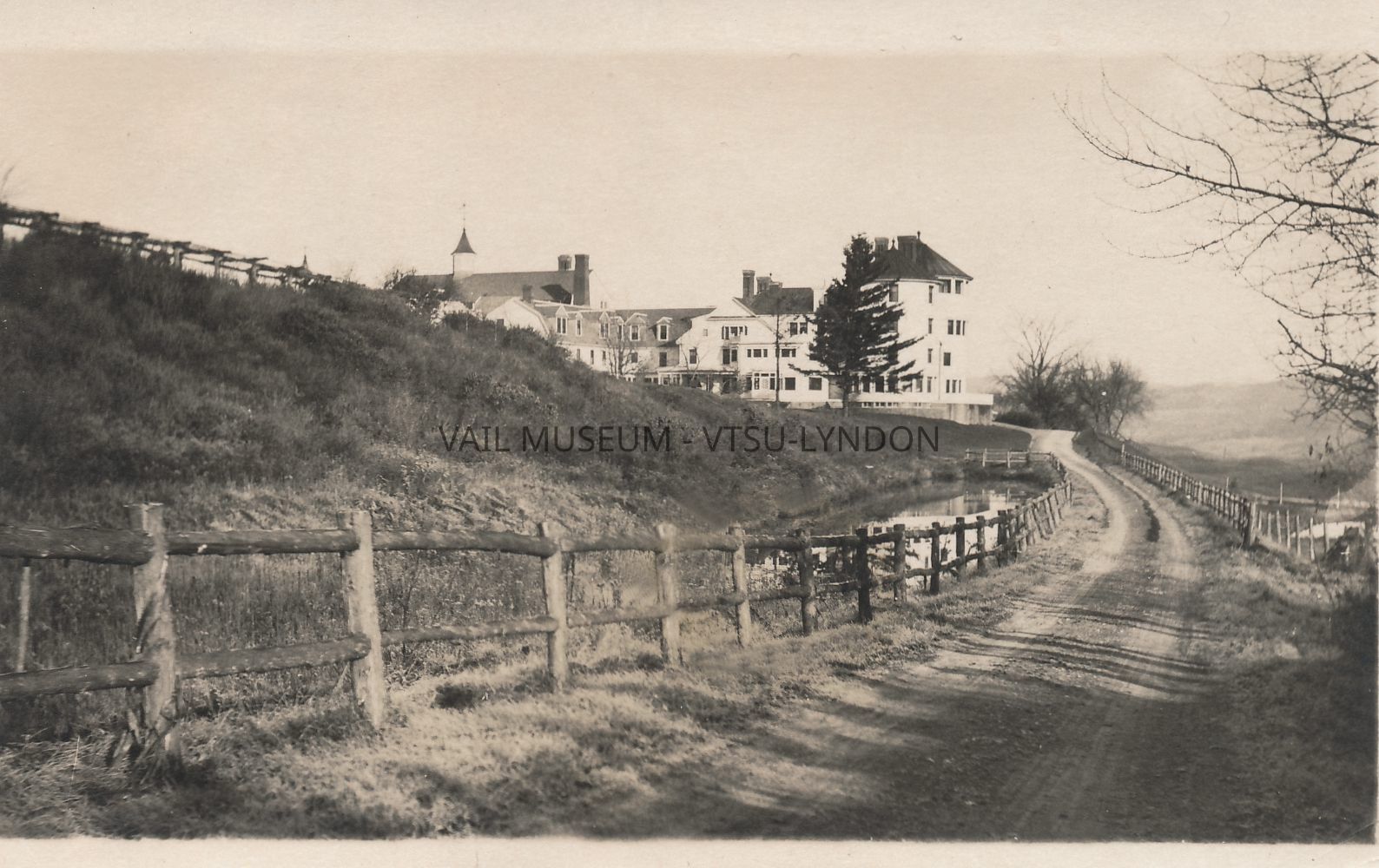 Vail Mansion showing the dirt road approach from the south with gates and fences for livestock, circa 1918: Real photo postcard - a portion of the trellis is visible in the upper left side of the photo - a small area of discoloration appears in the original digital image, lower center along the fence, roughly in the area of the fourth and fifth fencepost from the left