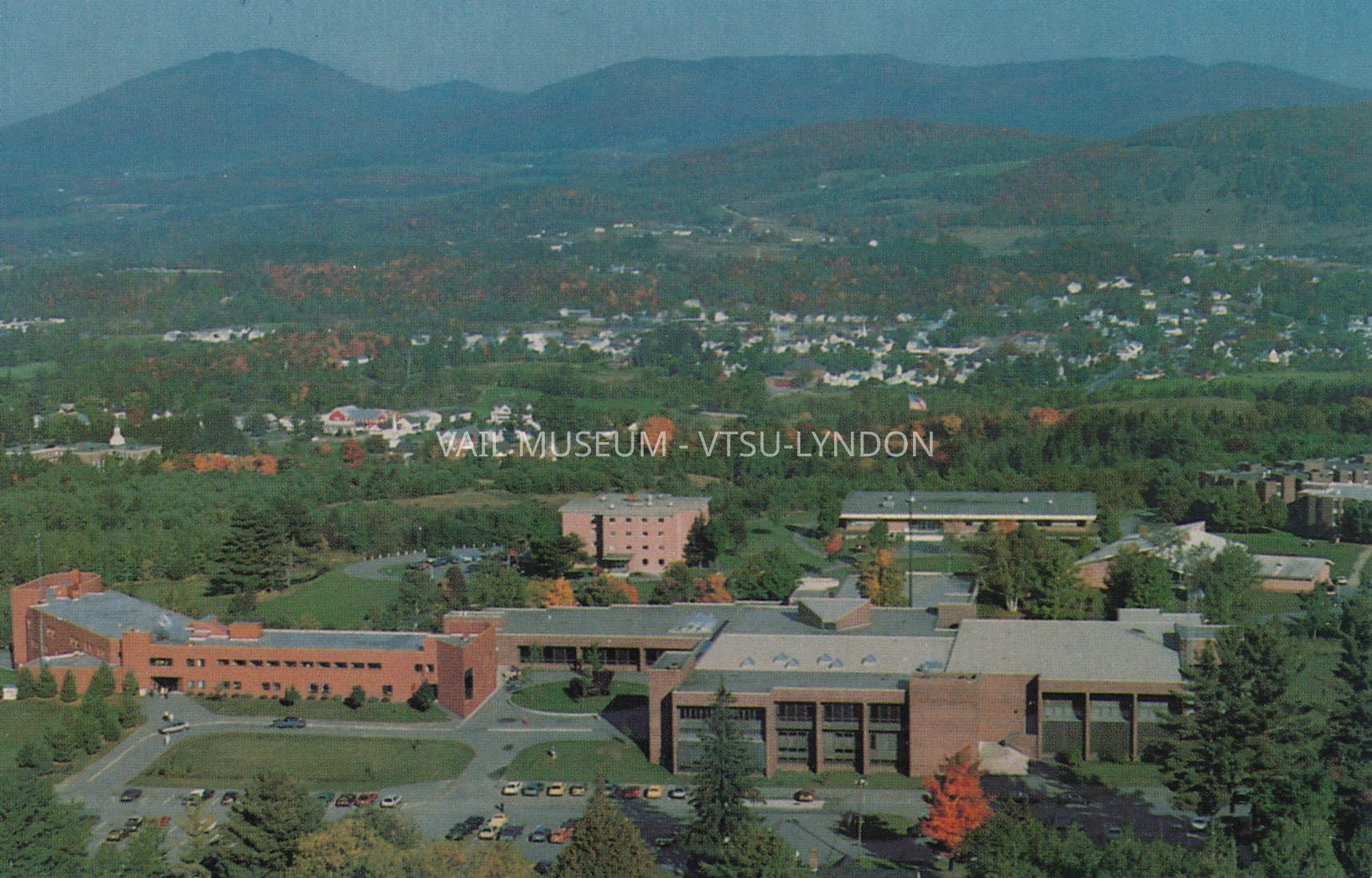 Color, aerial photo of Lyndon State College campus, looking east, circa 1990s, issued by Admissions, LSC.: From left, clockwise, T.N. Vail Center, Wheelock dormitory, Harvey Academic Center, Stonehenge dormitory complex, Stevens Dining Hall, Stannard Gym, Alexander Twilight Theatre and Fairbanks Science Wing. Samuel Read Hall Library roof is visible between the Alexander Twilight Theatre in the foreground and Harvey Academic Center at the far side, center right of the photo. A US flag flies atop the original flagpole from the T.N. Vail estate.