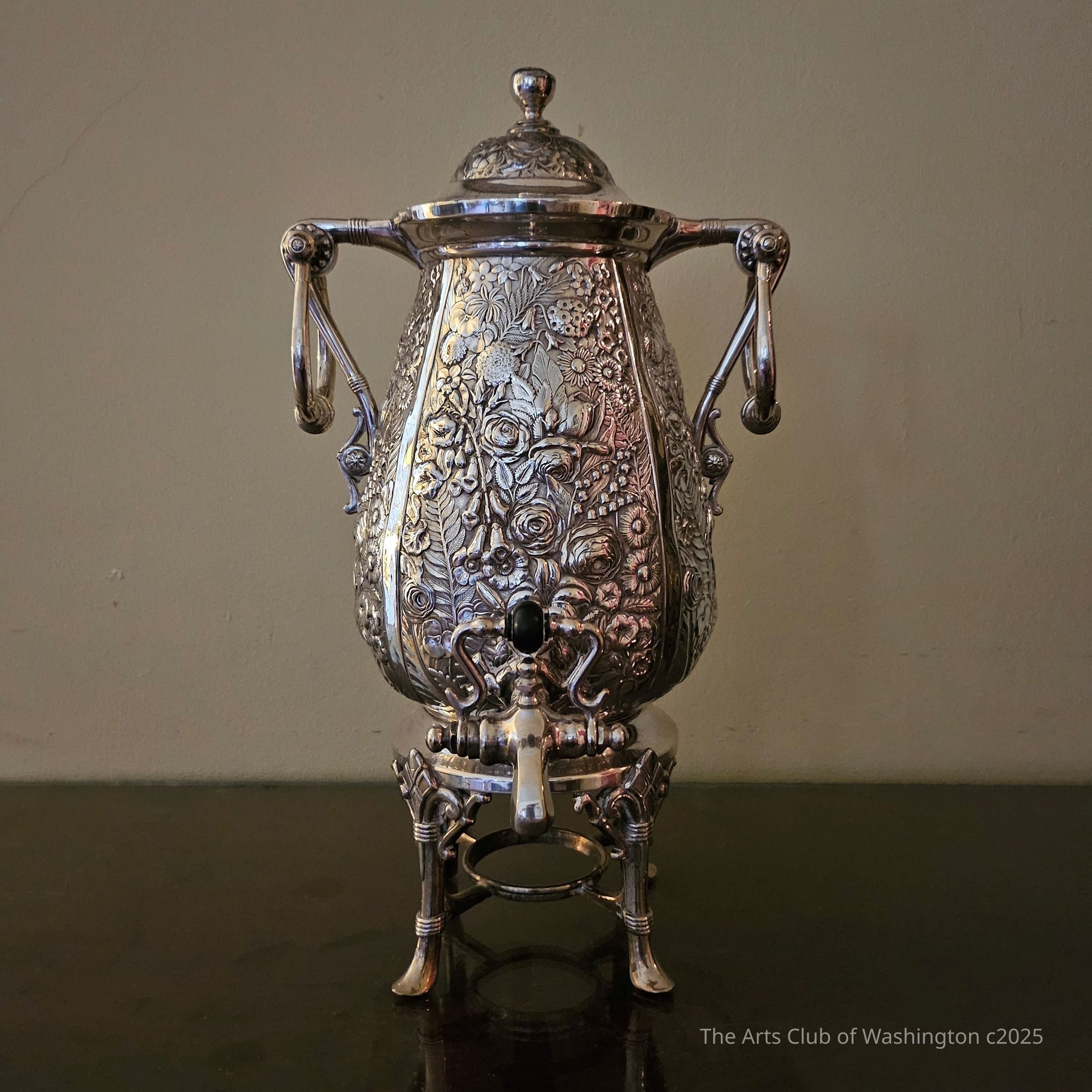 Urn, Coffee, Reed and Barton, silverplated metal, 1876.: Four straight bound and scrolled legs with slipper feet connect to a central fuel holder and support a bulbous lidded body heavily embossed with roses and other flowers and having a spigot with a handle formed by opposing "S" shapes. A pair of "akimbo" arms with elongated rings attach to each side.