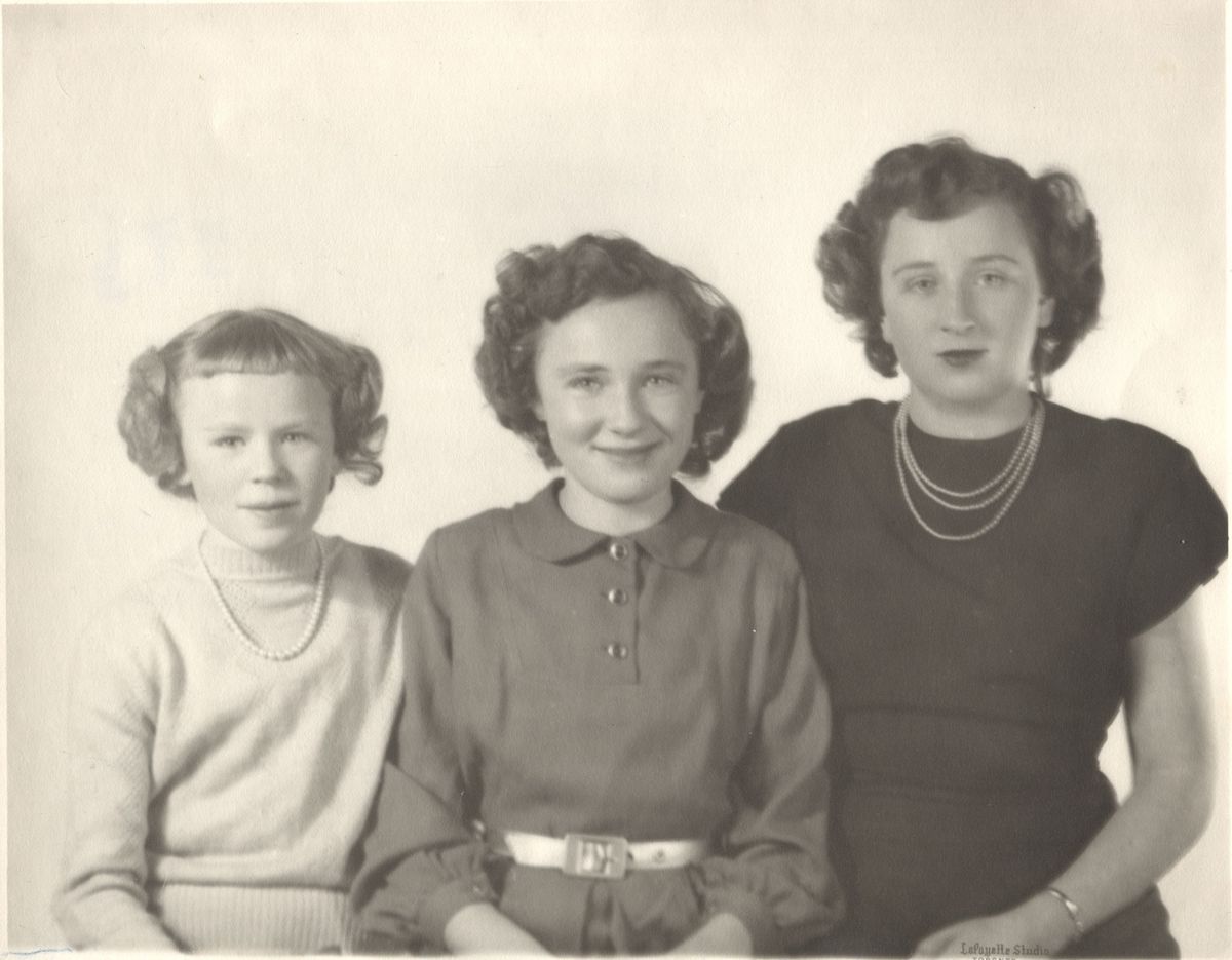 The Brown Sisters sat posing together for a portrait.: In this black-and-white photograph, three young women are seated together, looking directly at the camera with smiles. The woman on the left has a short haircut with bangs and wears a light-colored sweater adorned with a string of pearls. The woman in the middle is wearing a collared dress with a bely, and the last woman is wearing a black dress with a string of pearls.