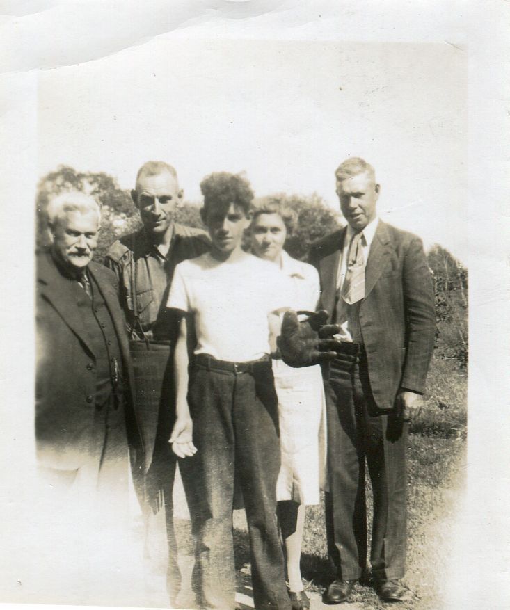 Shuter Haskill, Harry, Bill, and Mabel Wade, and Sid Haskill posing outside for a photo.: This is a faded black and white photo that has been cut at the bottom of five people standing together in a field with trees in the background. The first man on the left is wearing a dark coloured three piece suit and the man next to him is in trousers with suspenders over a collared shirt. The third person is a young boy who is standing slightly in front of everyone else and is wearing a white t-short with pants held up by a belt, in his left hand he is wearing an old school baseball glove. Behind him is a woman in a white collared dress and beside her is a man in a suit with a wide tie.