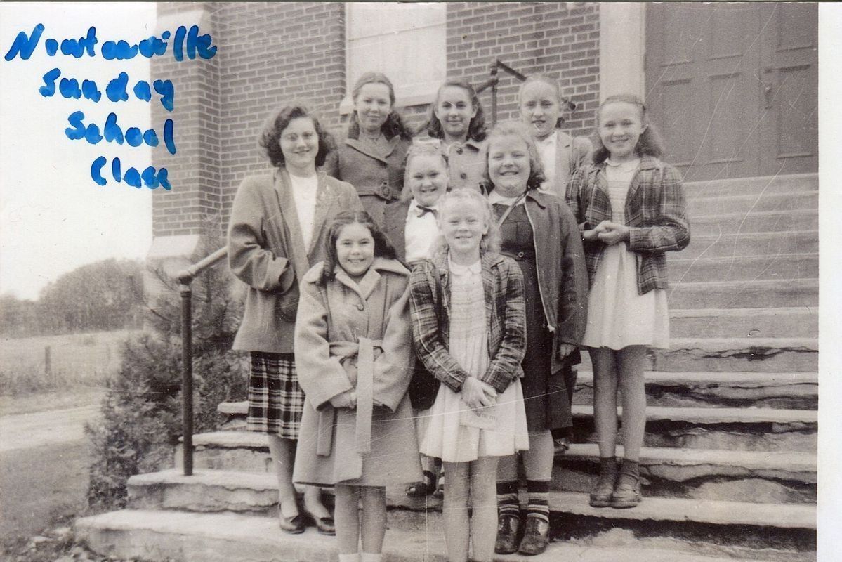 The Newtonville Sunday school class posing together on the steps for a photo.: The image shows a group of smiling young girls and their teacher, gathered on the steps of a brick church, for a photo labeled 'Newtonville Sunday School Class' in blue handwriting. The girls are dressed in coats and skirts, smiling at the camera, while standing holding their hands together in front of themselves. There's a collection of trees in the background, behind the fields.